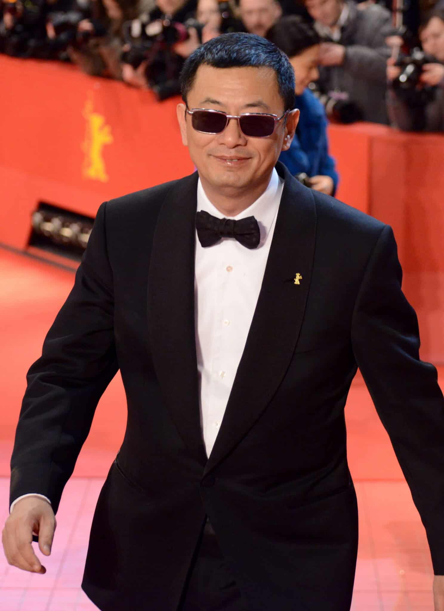 A man in a tuxedo and sunglasses smiles on a red carpet with photographers in the background.