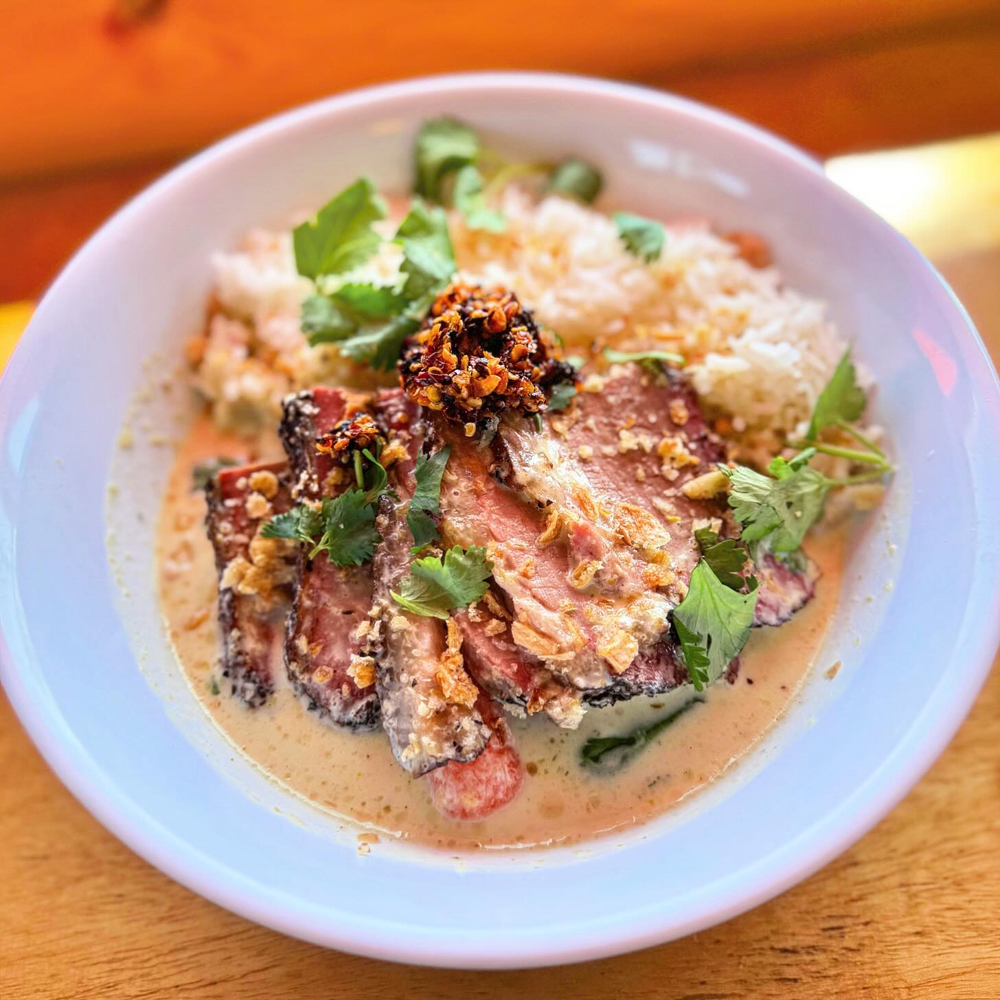 A bowl of creamy curry with sliced steak, white rice, carrots, cilantro, and chili oil, served in a white bowl on a wooden table.