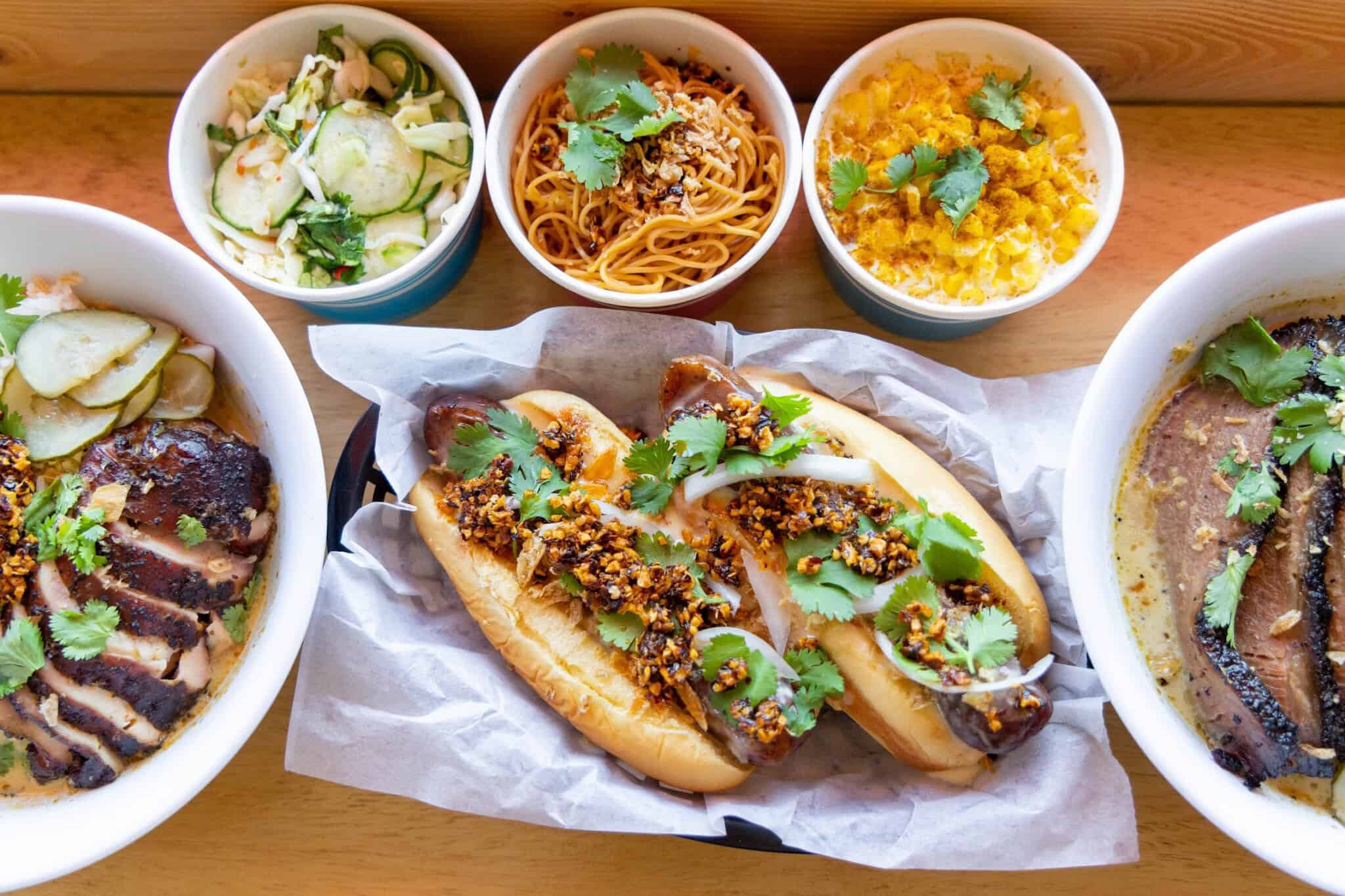 A top-down view of various dishes including grilled chicken, noodles, two hot dogs topped with onions and herbs, cucumber salad, and a baked dish, all garnished with fresh cilantro.