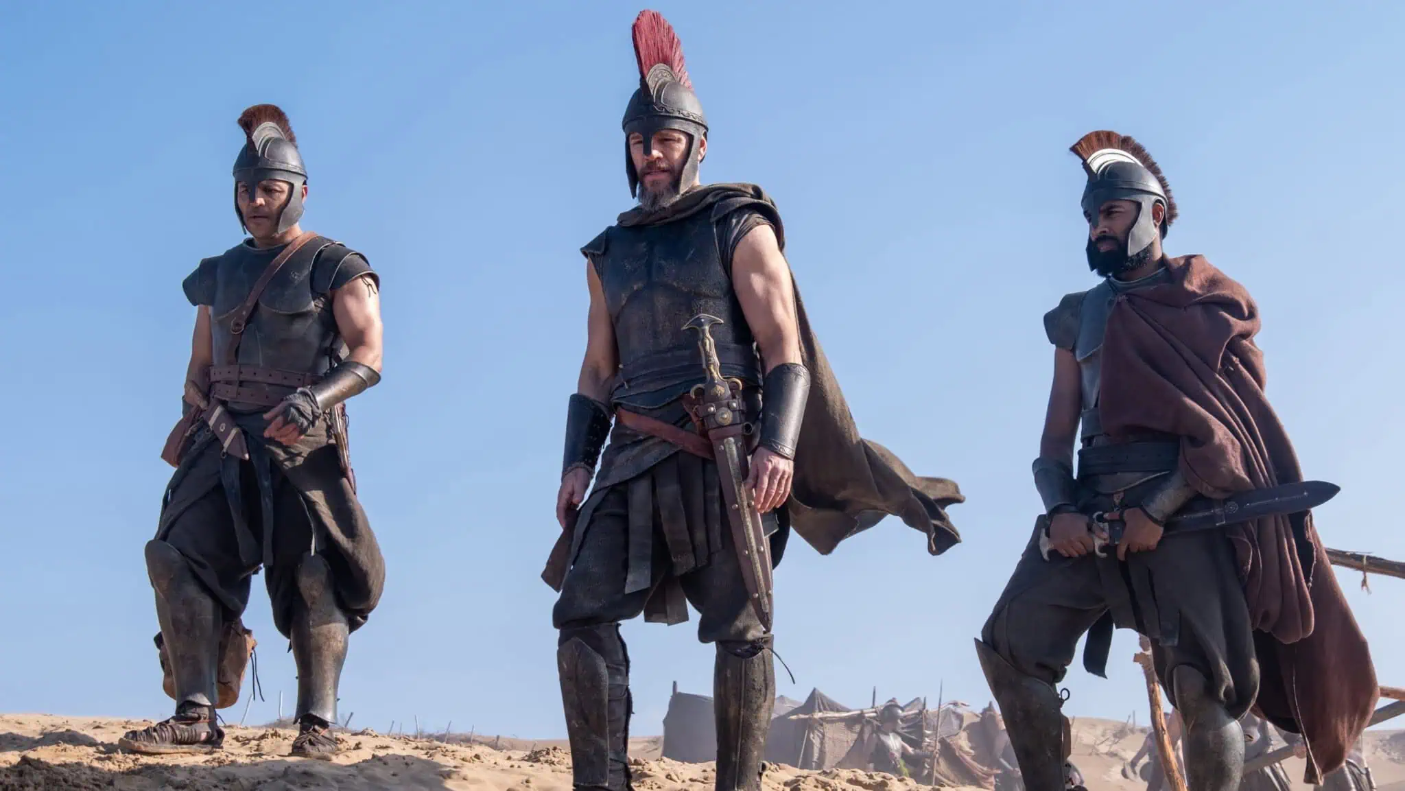 Three men dressed as ancient warriors in armor and helmets stand on sandy ground under a clear blue sky, looking serious and alert, with tents visible in the background.