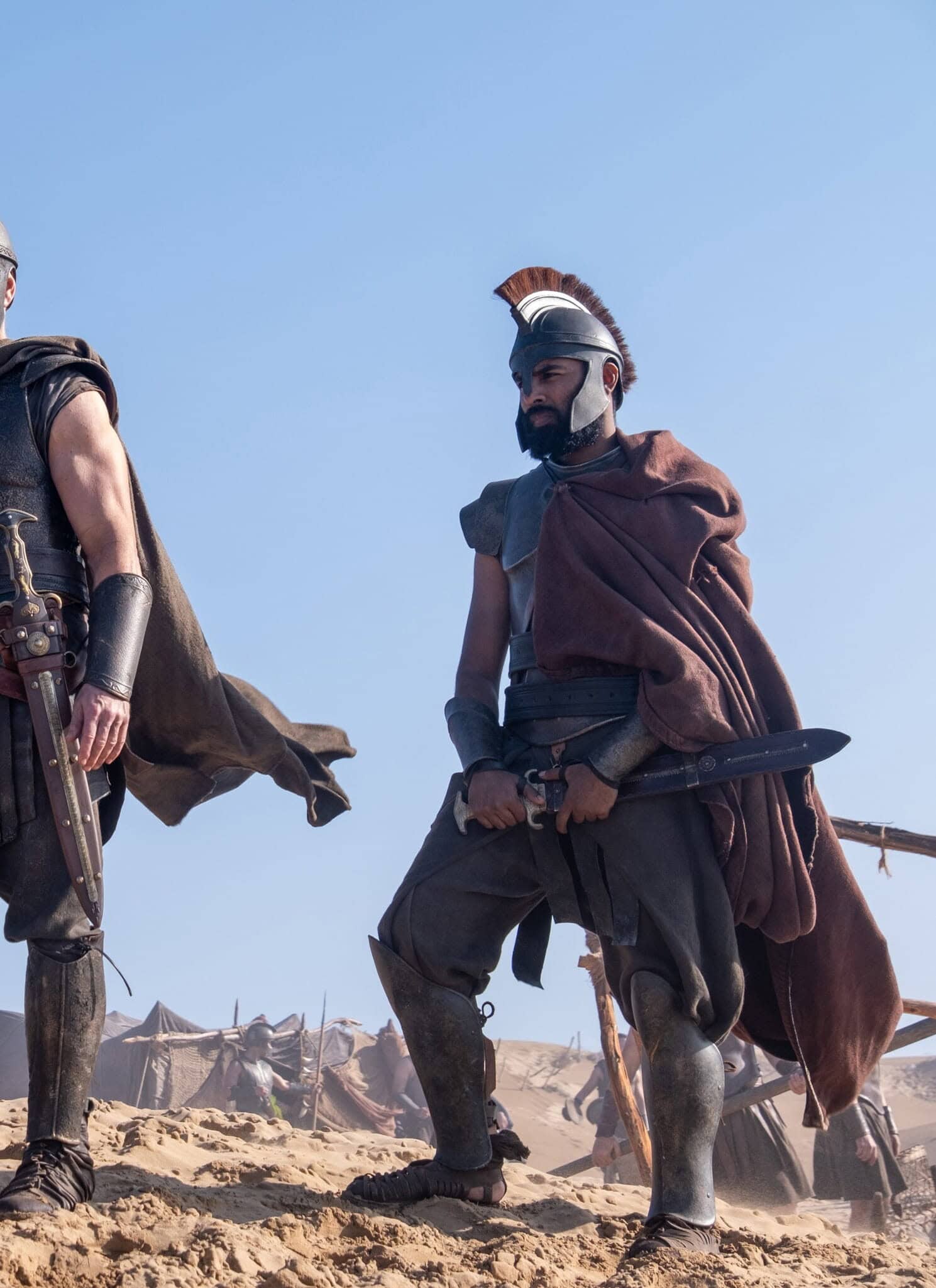 Three men dressed as ancient warriors in armor and helmets stand on sandy ground under a clear blue sky, looking serious and alert, with tents visible in the background.