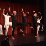 Six dancers in colorful turbans and traditional outfits perform a synchronized dance on stage in front of a red curtain, each with one leg lifted and arms raised.