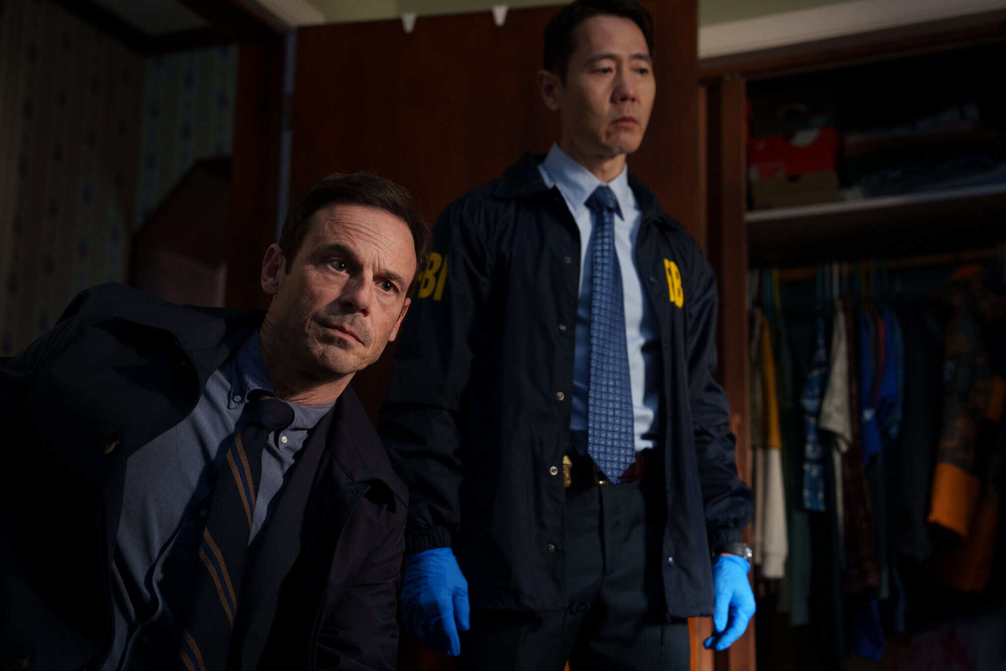 Two men in suits, wearing FBI jackets and blue gloves, stand in a dimly lit room near an open closet, looking intently at something off-frame. Clothing hangs in the background.