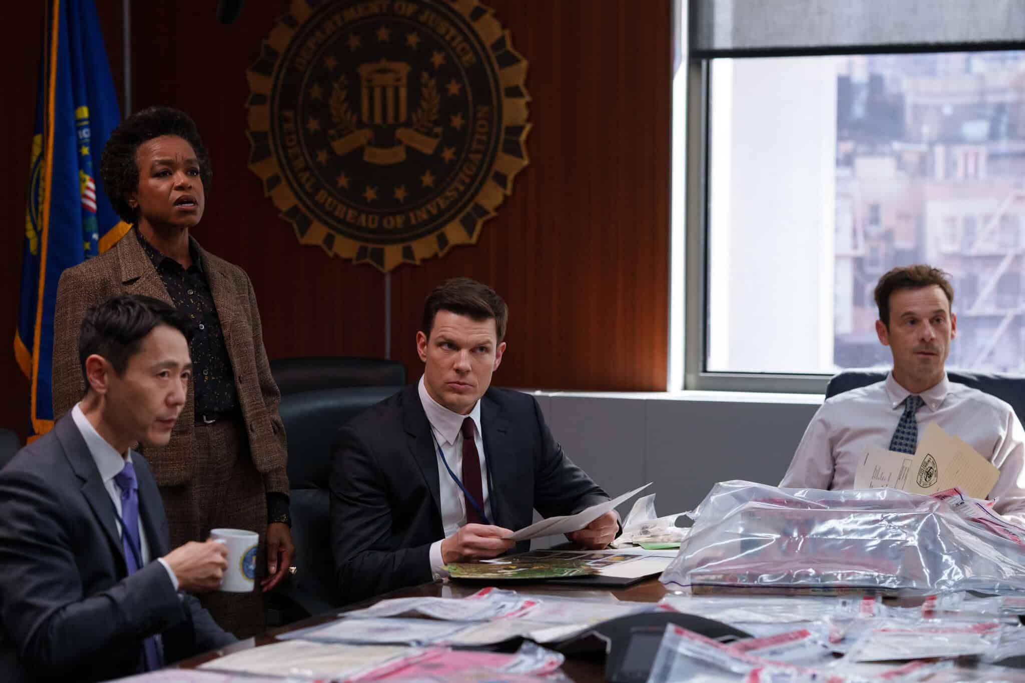 Four people in formal clothes sit and stand around a conference table covered in evidence bags and files, with a large FBI seal on the wall behind them.