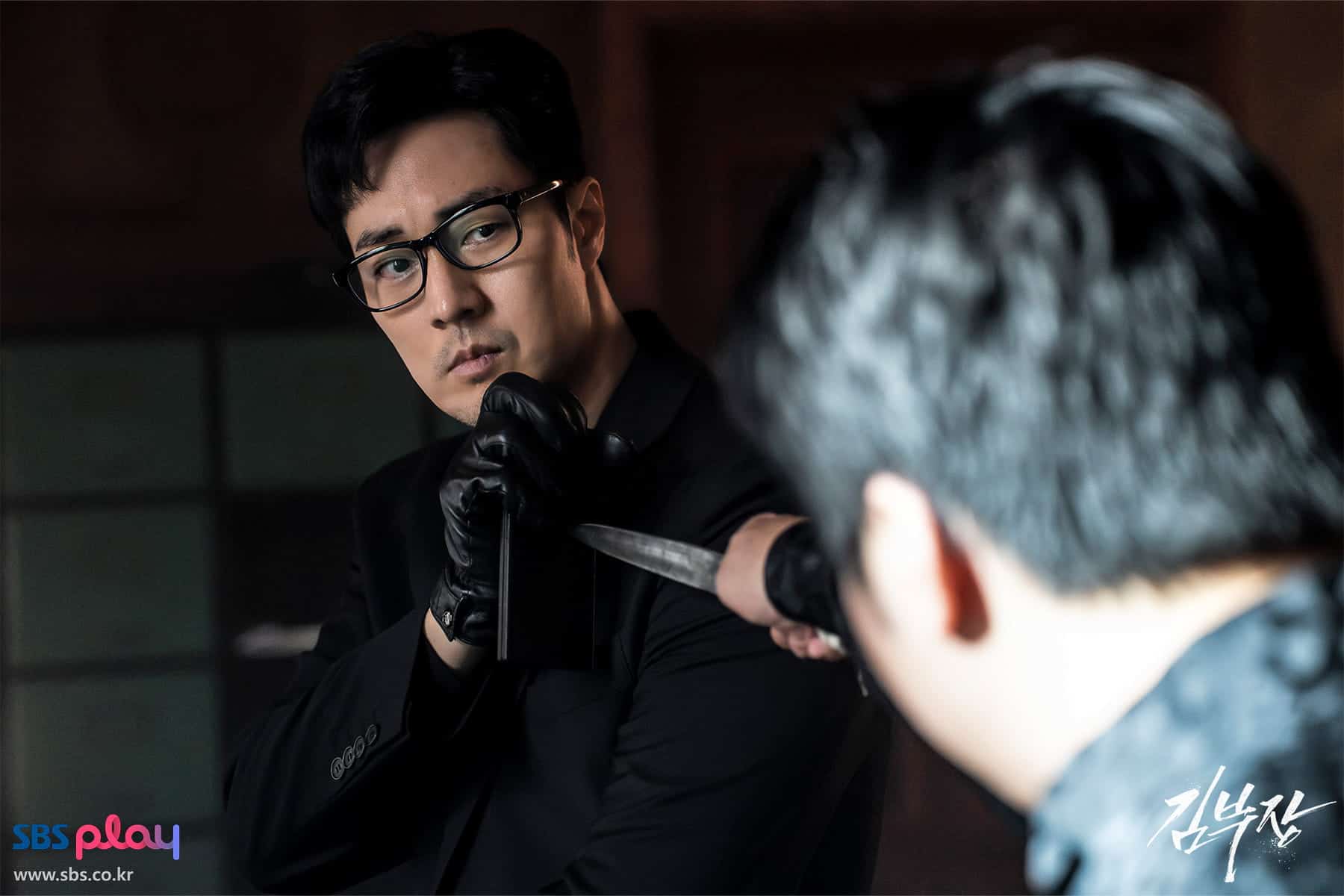 A man in black with glasses and gloves holds another mans arm, stopping a knife attack. The scene is intense and dramatic, with both men focused on each other. SBS play and Korean text are visible in the bottom corners.