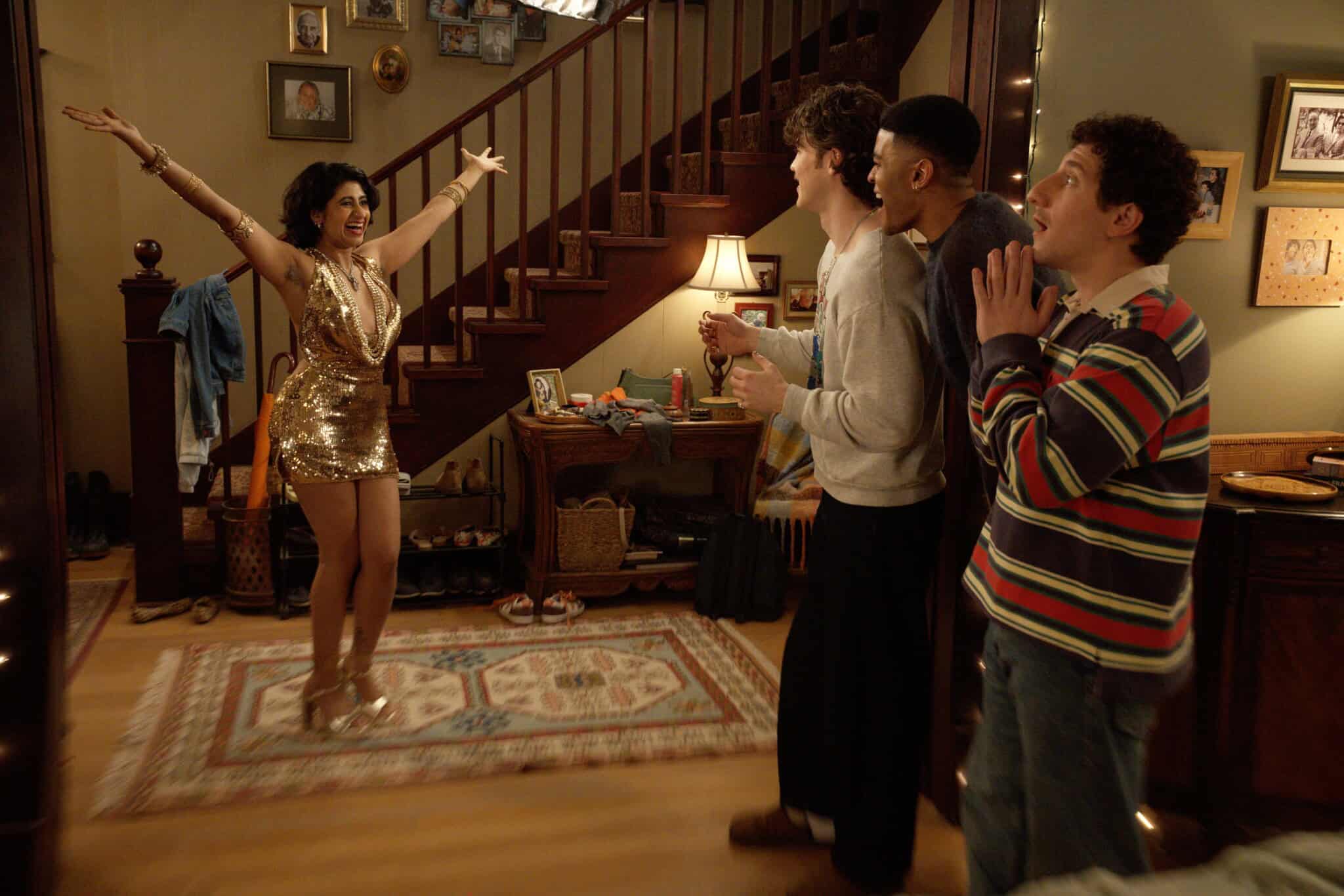 Amita Rao, dressed in a sparkling gold gown, poses confidently with arms outstretched as three surprised men stand near a staircase, reacting with excitement and amazement in a cozy, warmly lit living room.