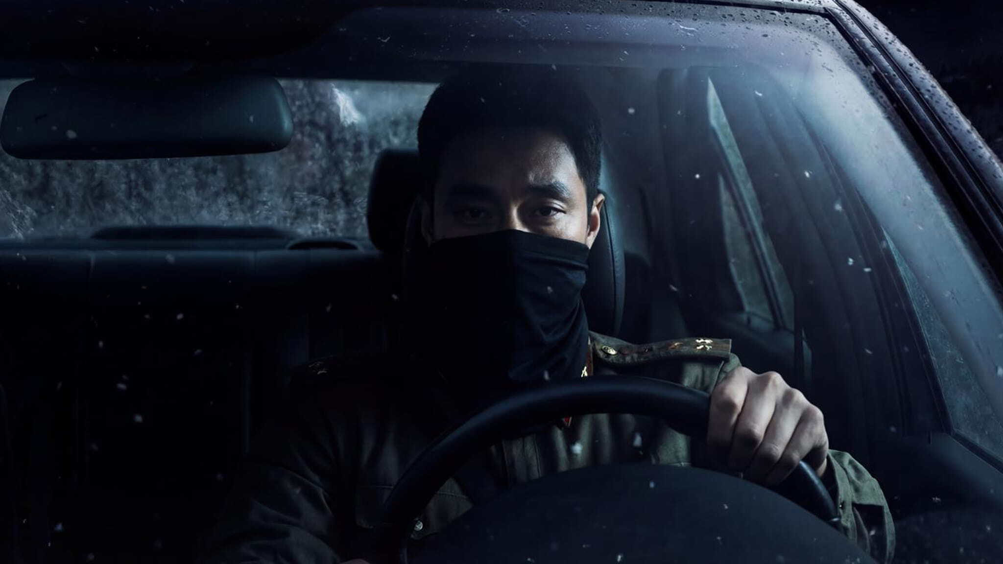 A person wearing a black face covering sits behind the wheel of a car at night, gripping the steering wheel and looking forward through a rain-speckled windshield.