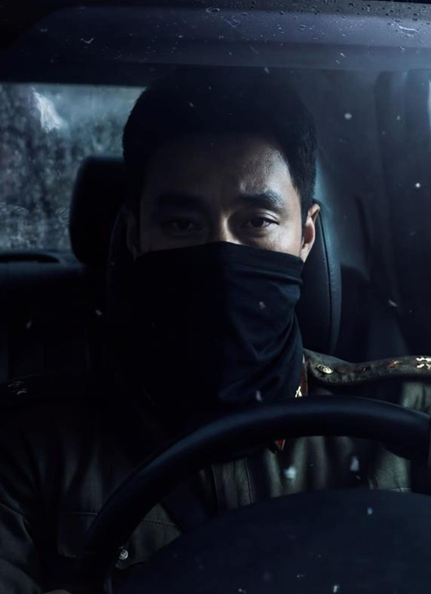 A person wearing a black face covering sits behind the wheel of a car at night, gripping the steering wheel and looking forward through a rain-speckled windshield.