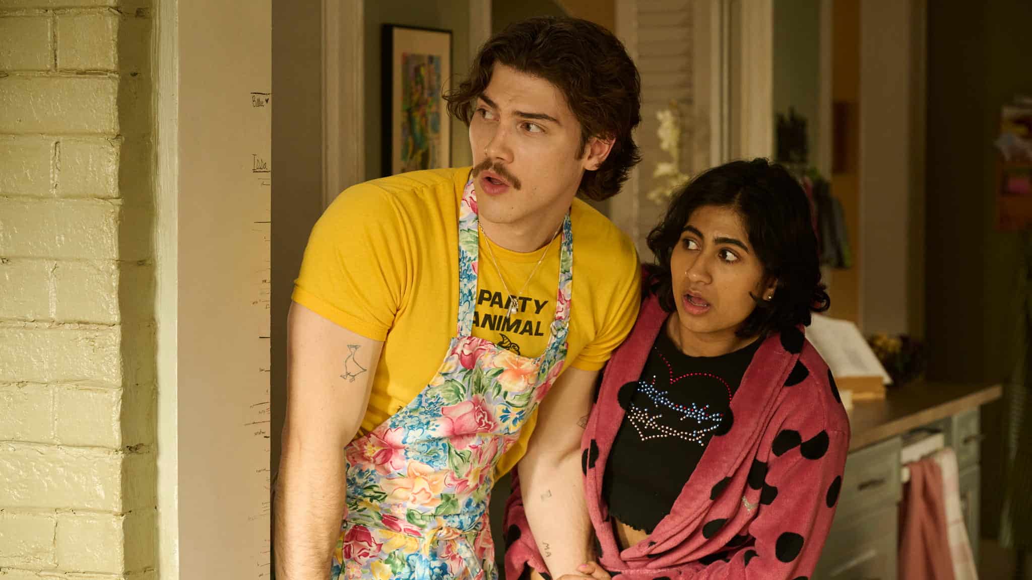 A young man in a floral apron and yellow shirt stands next to a surprised young woman, Amita Rao, in a pink polka-dot robe. Both look off to the left, appearing shocked or concerned.