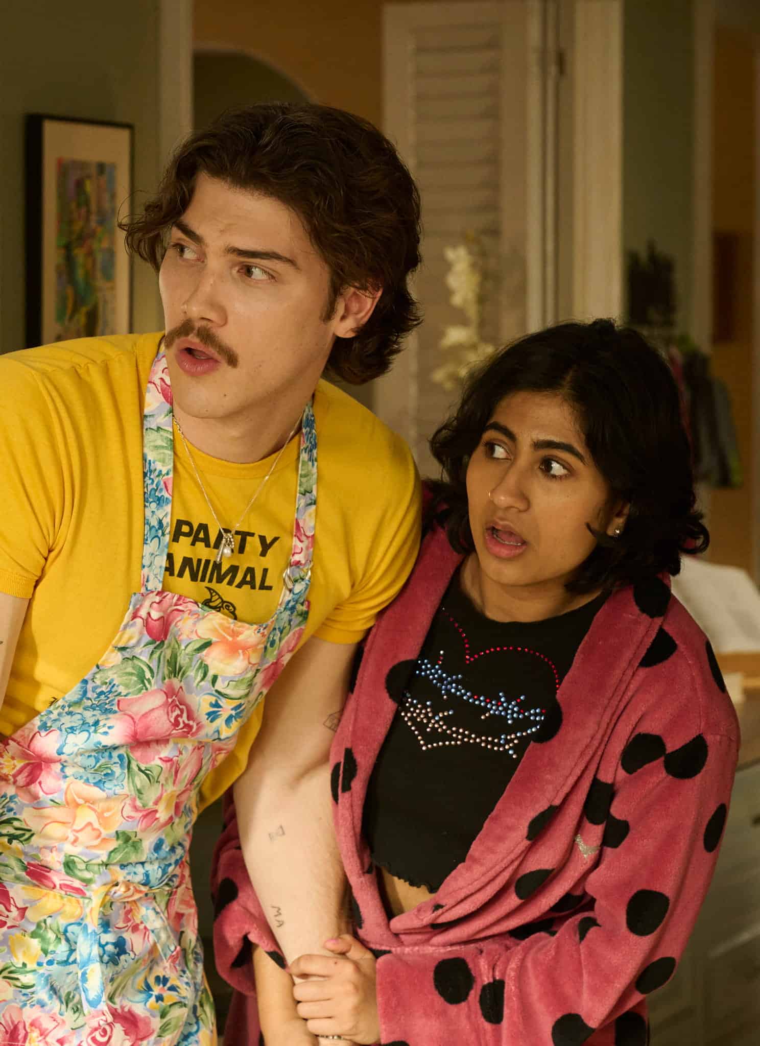 A young man in a floral apron and yellow shirt stands next to a surprised young woman, Amita Rao, in a pink polka-dot robe. Both look off to the left, appearing shocked or concerned.