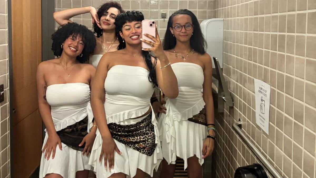 Four women in matching white dresses with leopard print sashes pose for a mirror selfie in a tiled bathroom. Three smile, while one stands with her hand raised behind her head.