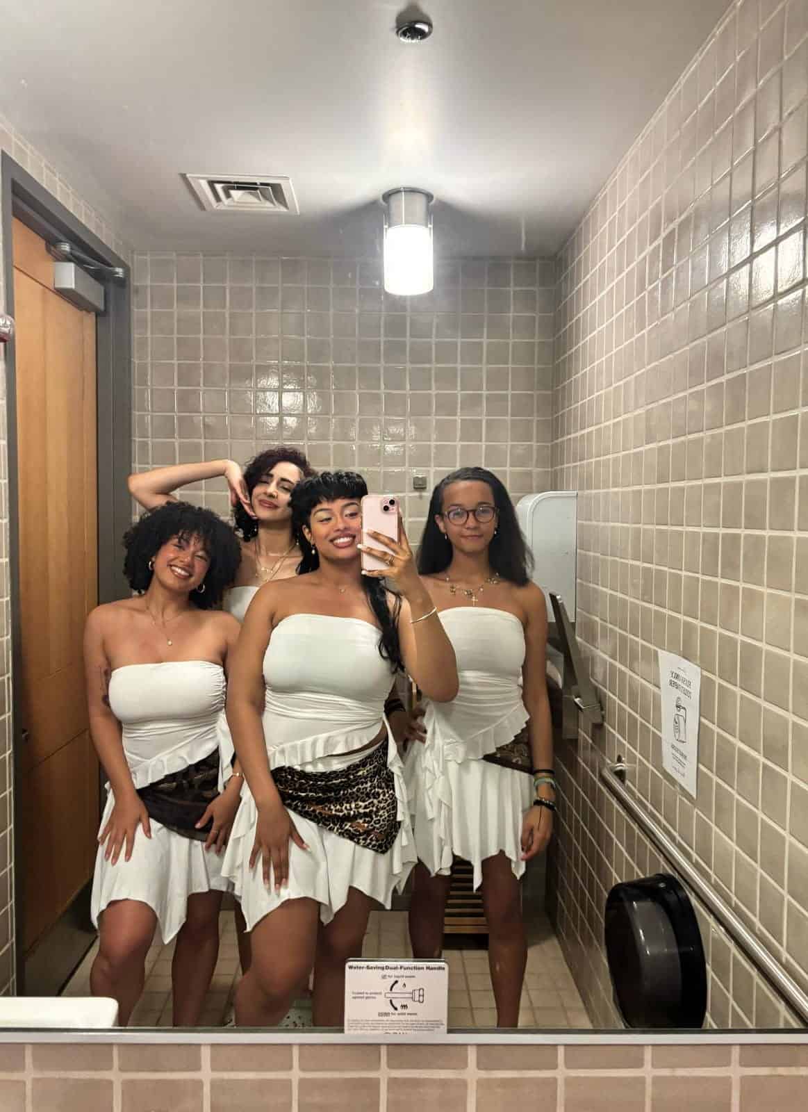 Four women in matching white dresses with leopard print sashes pose for a mirror selfie in a tiled bathroom. Three smile, while one stands with her hand raised behind her head.