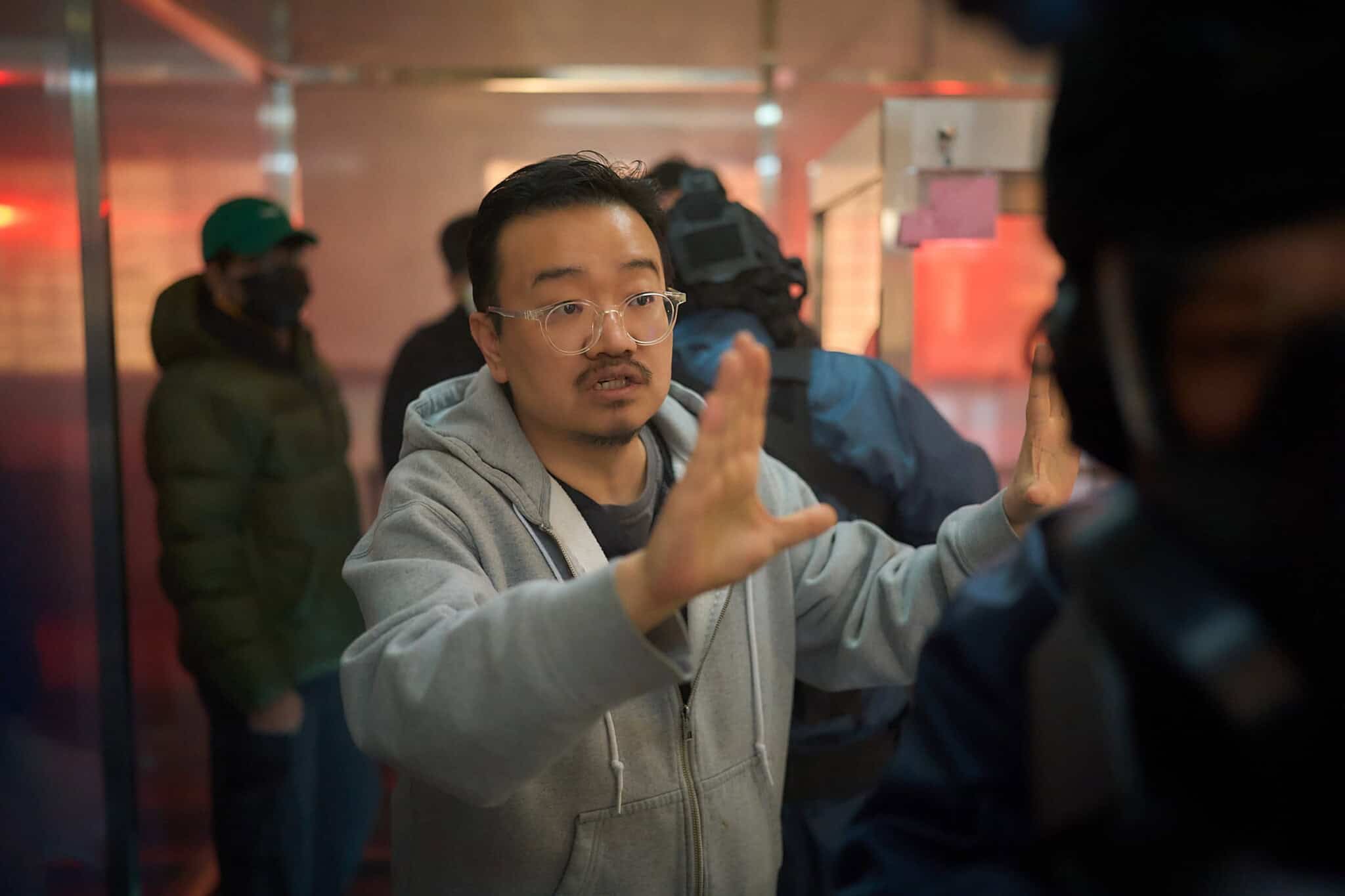 A man in glasses and a gray hoodie gestures with his hands, appearing to give directions in a room with several people—some in tactical gear—all set against a red glow.