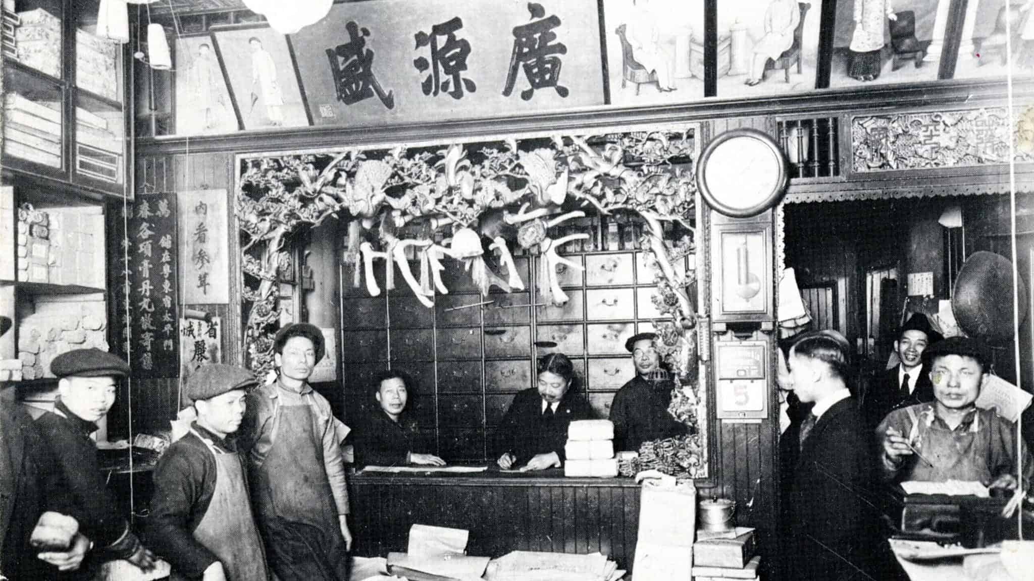 Black-and-white photo of an old Chinese apothecary with staff and customers. Shelves, drawers, herbs, and hanging dried goods surround the busy counter. Chinese characters are displayed on signs and banners.