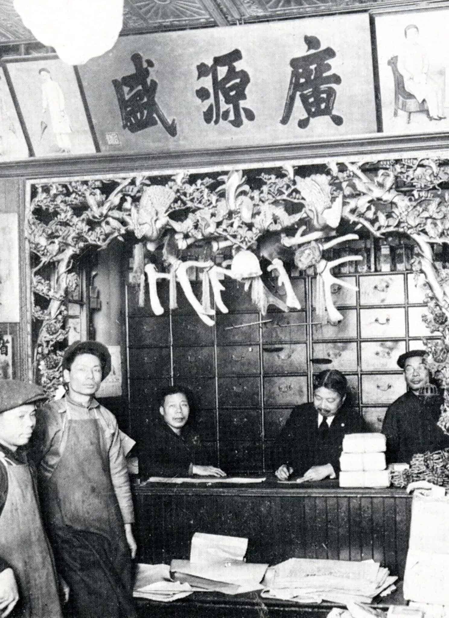Black-and-white photo of an old Chinese apothecary with staff and customers. Shelves, drawers, herbs, and hanging dried goods surround the busy counter. Chinese characters are displayed on signs and banners.