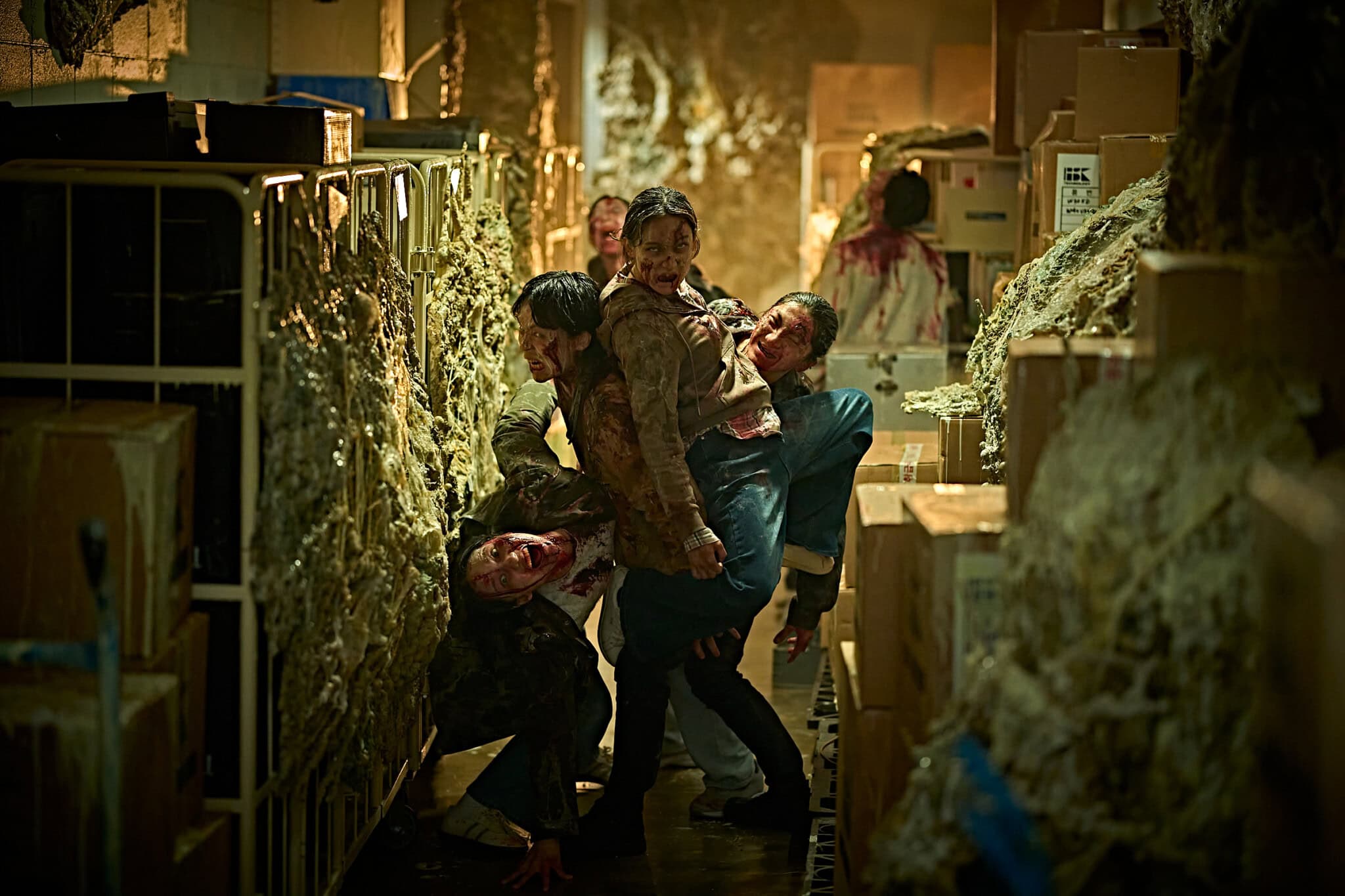 A woman fights off attacking zombies in a narrow, cluttered hallway lined with boxes and debris, fighting to protect her colony, while other survivors appear in the background.