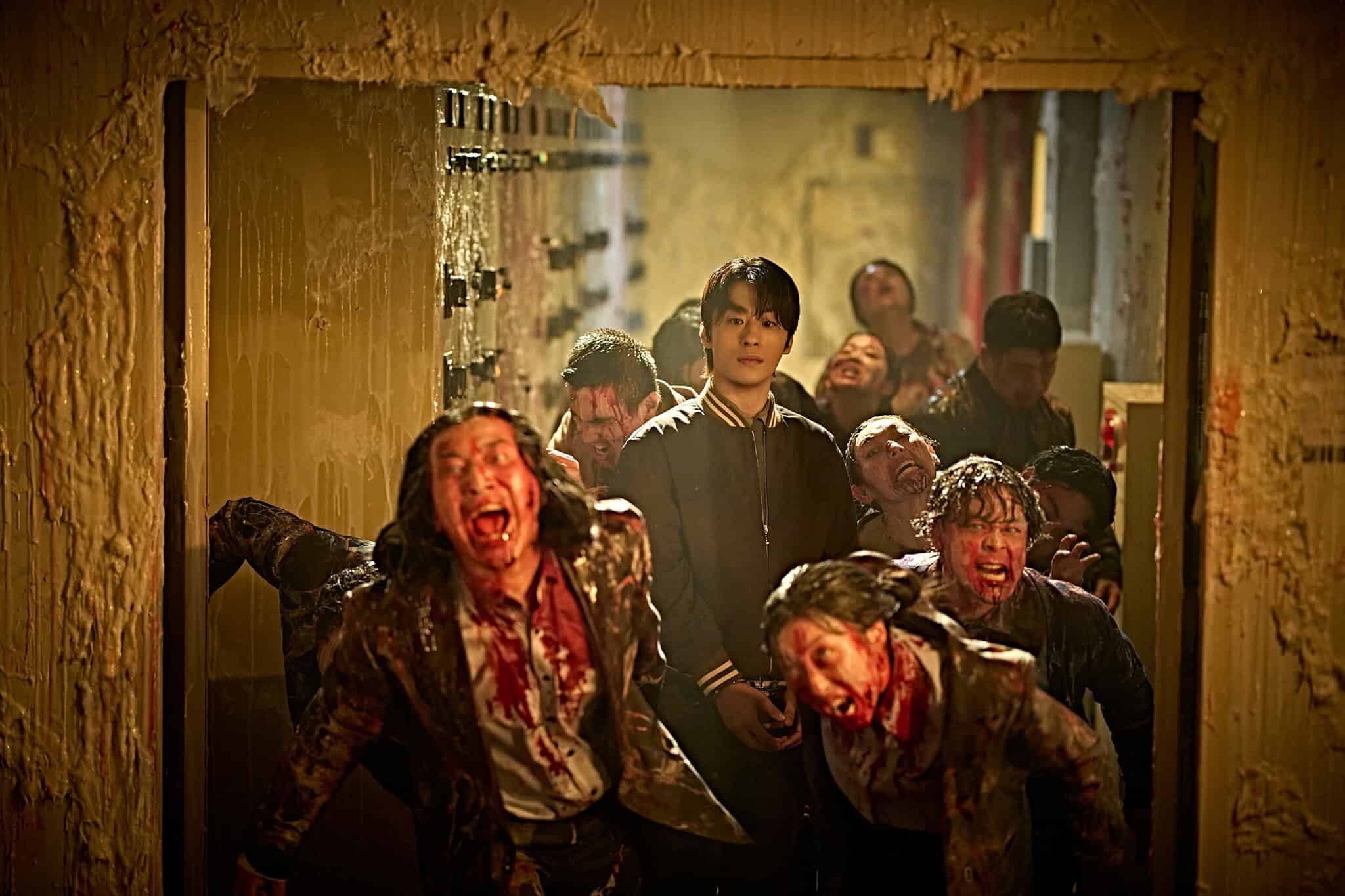A calm young man stands in a doorway, surrounded by blood-covered zombies lunging outward in a dimly lit, chaotic scene.