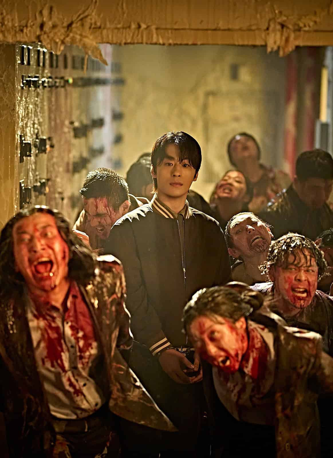A calm young man stands in a doorway, surrounded by blood-covered zombies lunging outward in a dimly lit, chaotic scene.