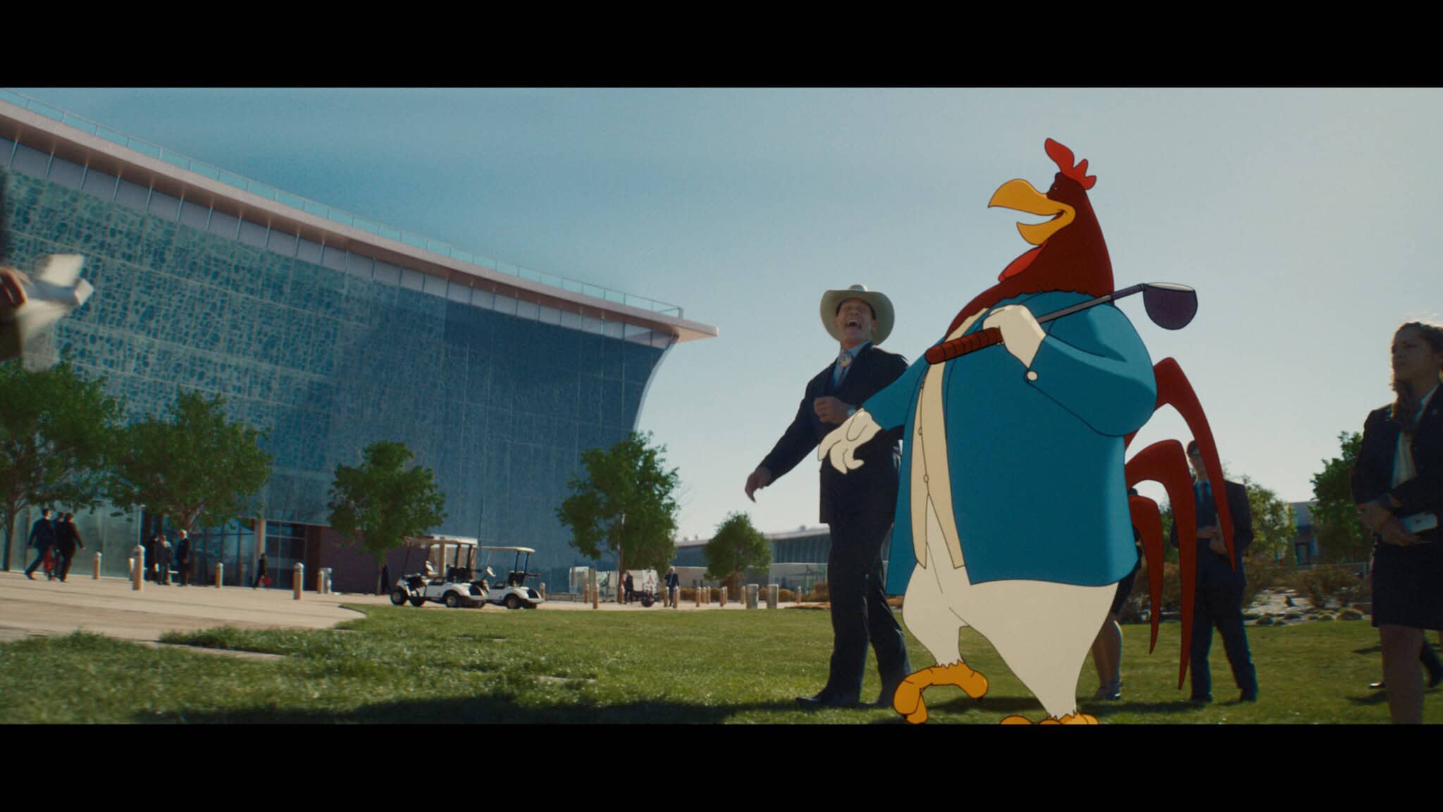 A man in a cowboy hat stands outside a modern building, looking up. Next to him is a large, animated rooster in a suit holding a golf club. Several other people are seen in the background.