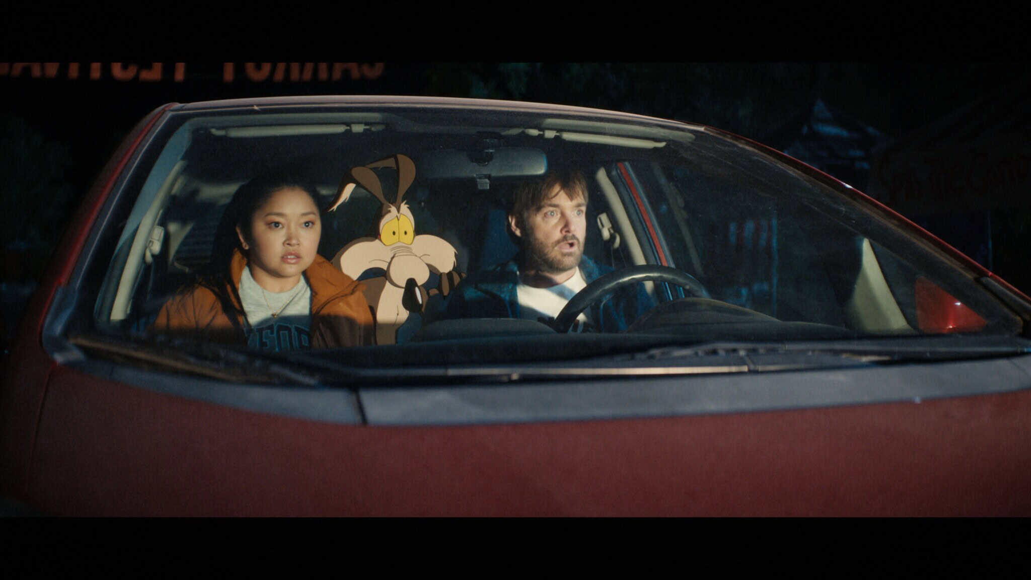 A young woman, a cartoon coyote, and a man sit together in the front seat of a red car at night, all looking surprised or shocked through the windshield.