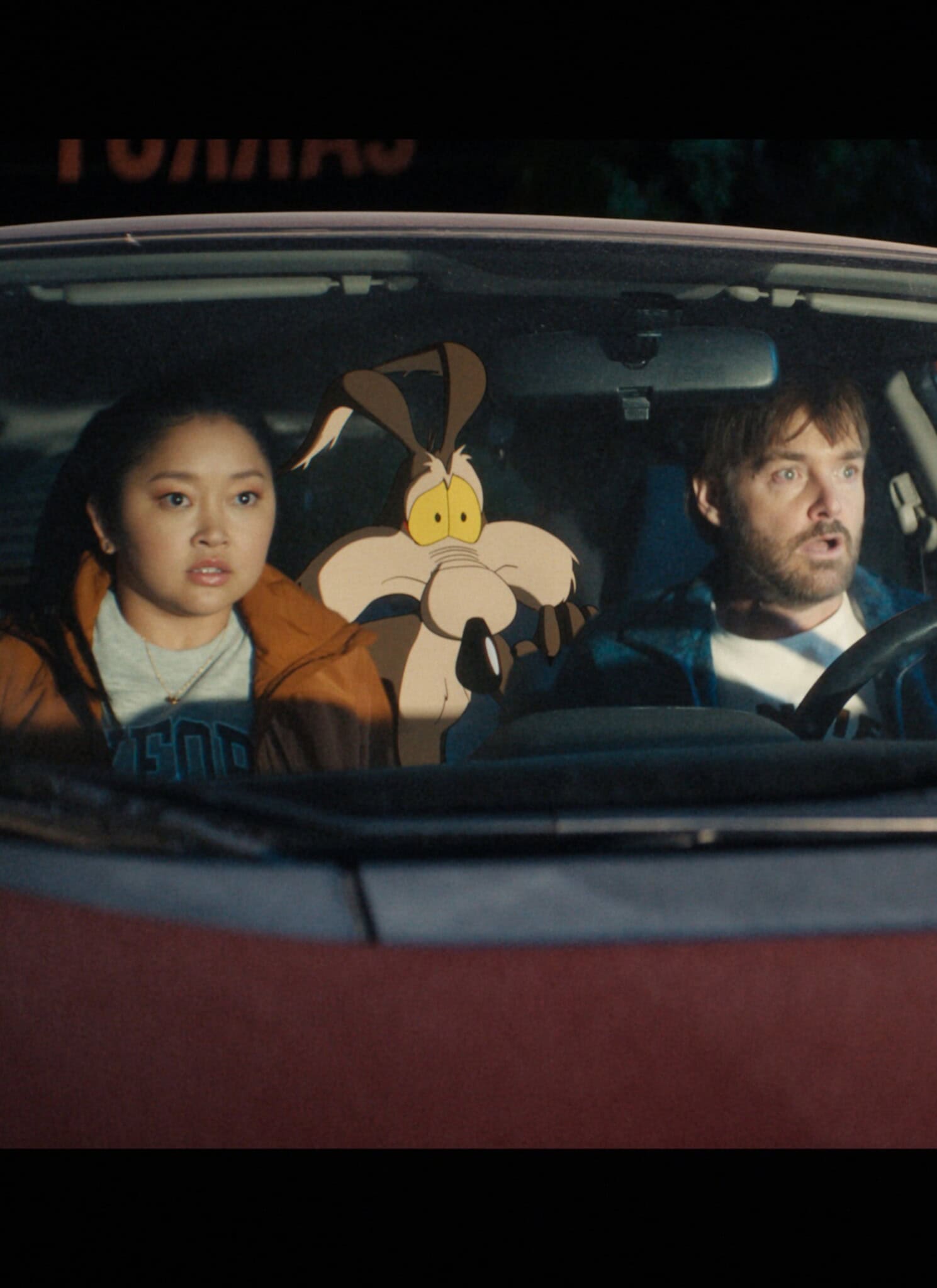 A young woman, a cartoon coyote, and a man sit together in the front seat of a red car at night, all looking surprised or shocked through the windshield.