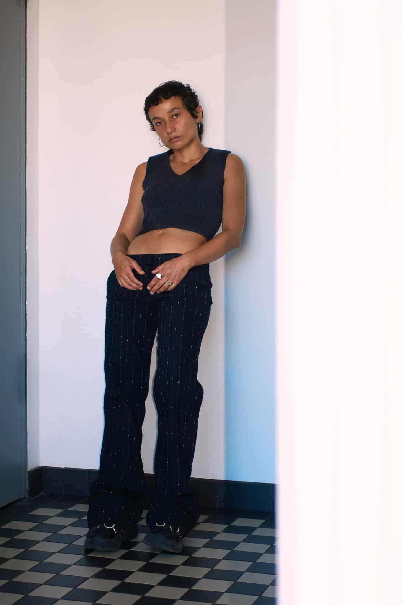 Person with short curly hair wears a sleeveless dark crop top and black pants with white pinstripes, standing against a wall on a black-and-white checkered floor, looking at the camera with a relaxed posture reminiscent of Nene H’s effortlessly cool style.