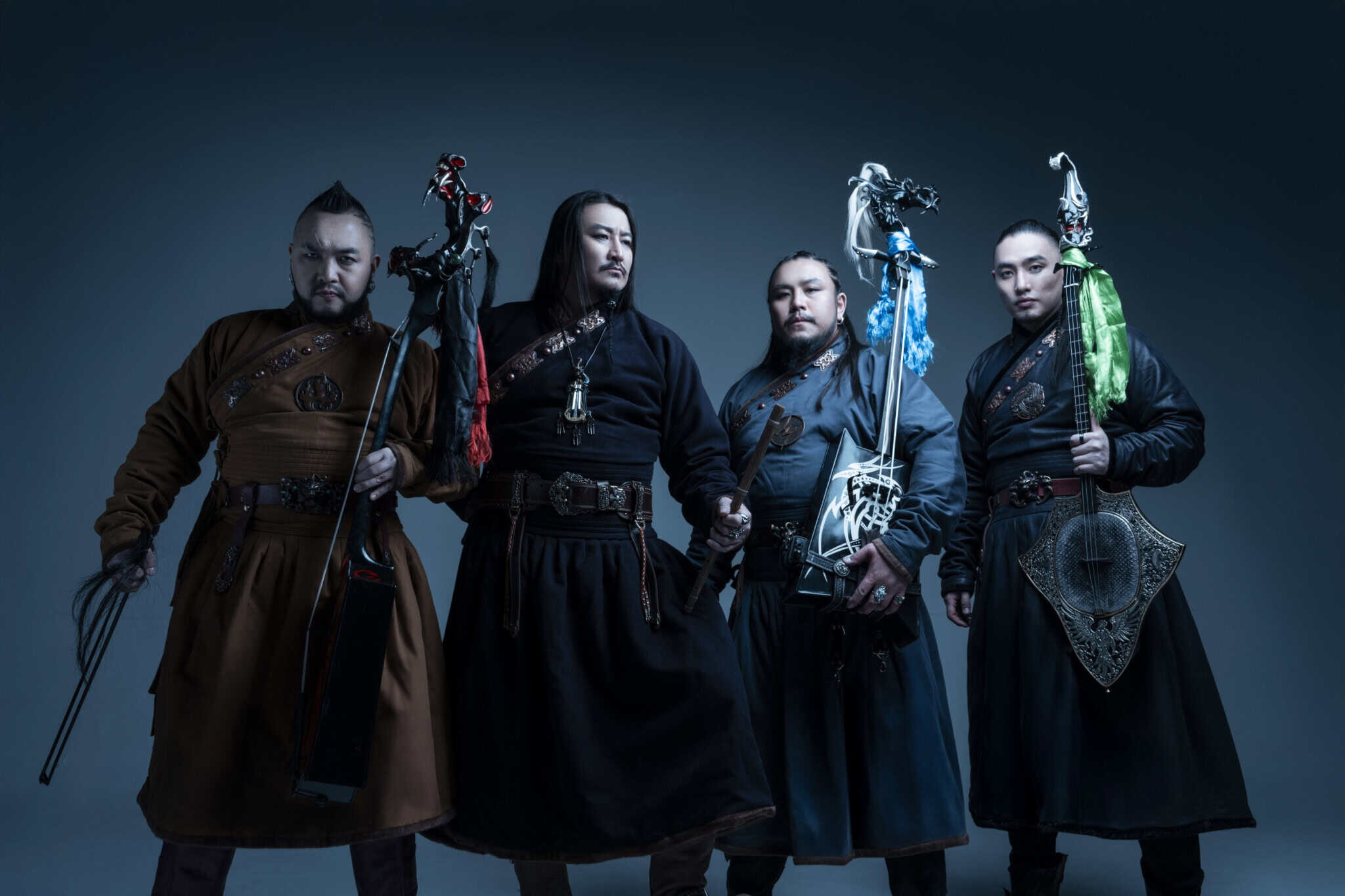 Four men in traditional Mongolian attire stand holding stringed instruments with decorative elements, facing forward against a dark, gradient background.