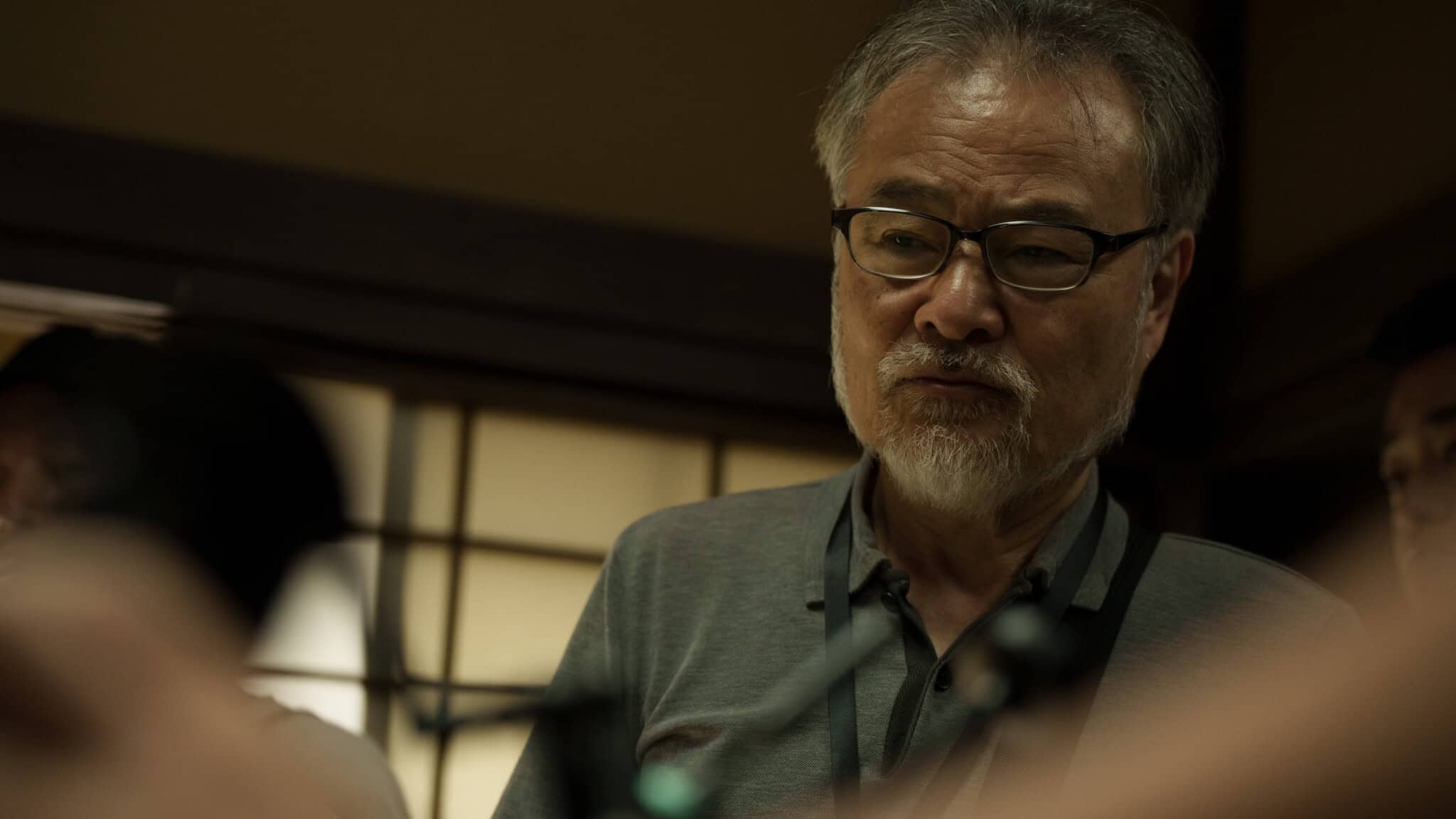 An older man with glasses and a gray beard looks down thoughtfully indoors, standing in soft, warm lighting with a shoji screen in the background.