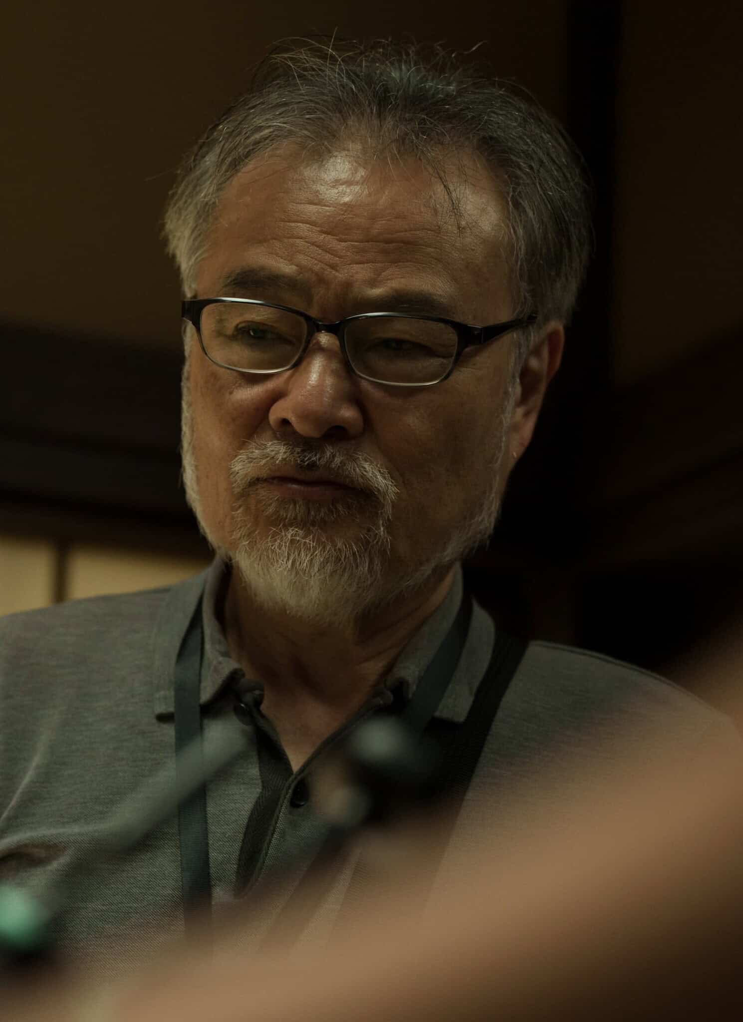 An older man with glasses and a gray beard looks down thoughtfully indoors, standing in soft, warm lighting with a shoji screen in the background.