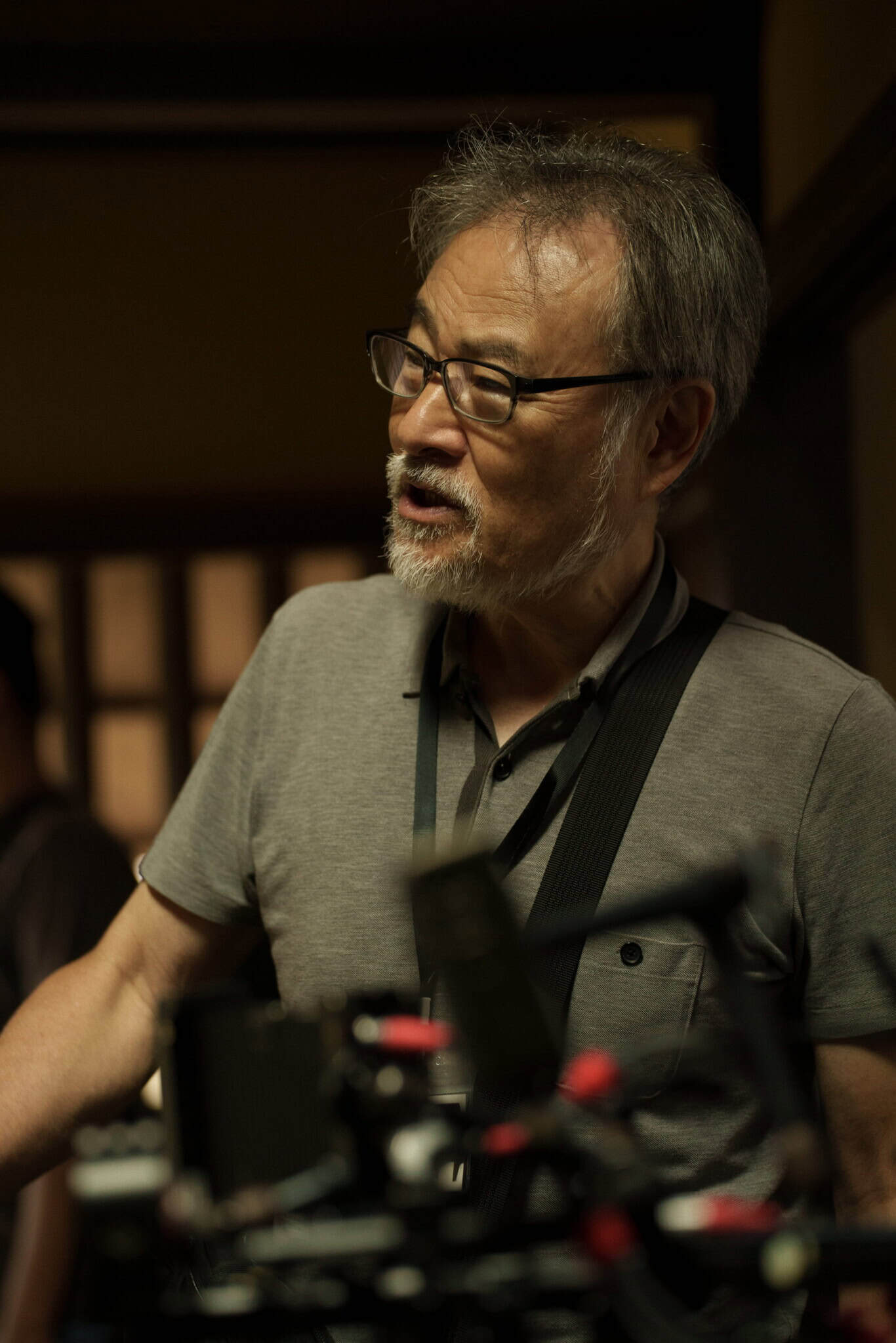 An older man with glasses and a gray beard stands indoors, wearing a gray polo shirt and camera strap, speaking near film equipment.
