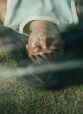 A person with long dark hair lies on grass with eyes closed, sunlight and shadows crossing their face, creating a serene and peaceful atmosphere.