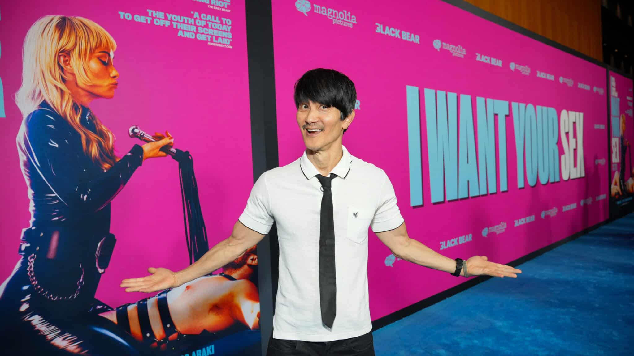 A man in a white shirt and black tie poses smiling with arms outstretched in front of a bright pink movie backdrop that reads I WANT YOUR SEX and features a woman holding a microphone.