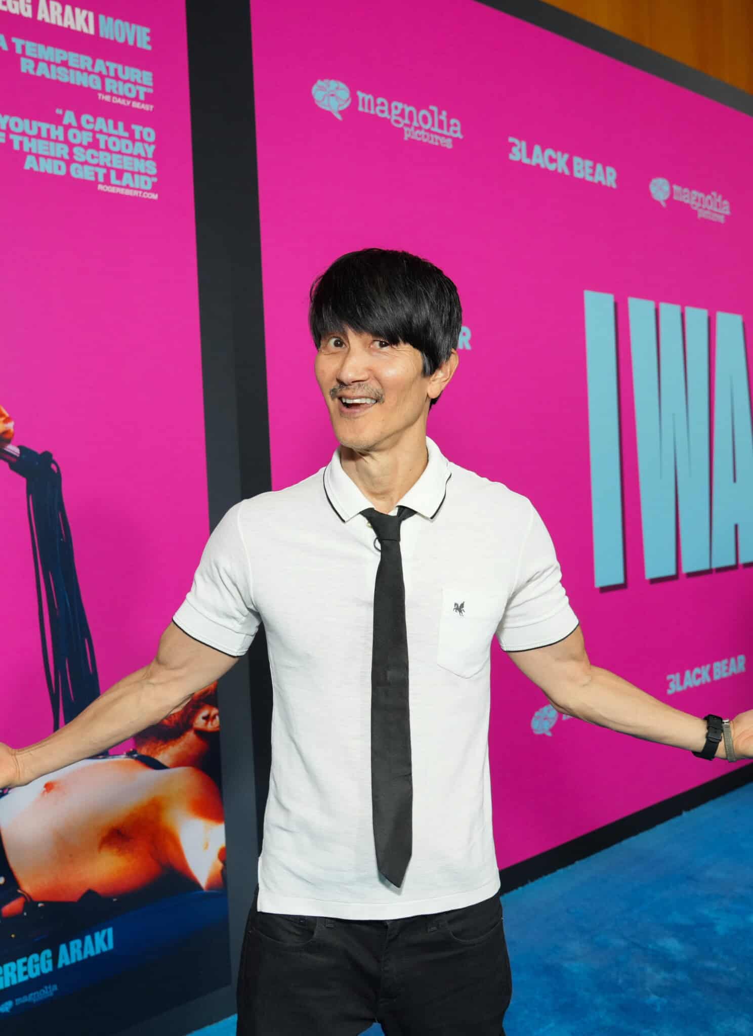 A man in a white shirt and black tie poses smiling with arms outstretched in front of a bright pink movie backdrop that reads I WANT YOUR SEX and features a woman holding a microphone.
