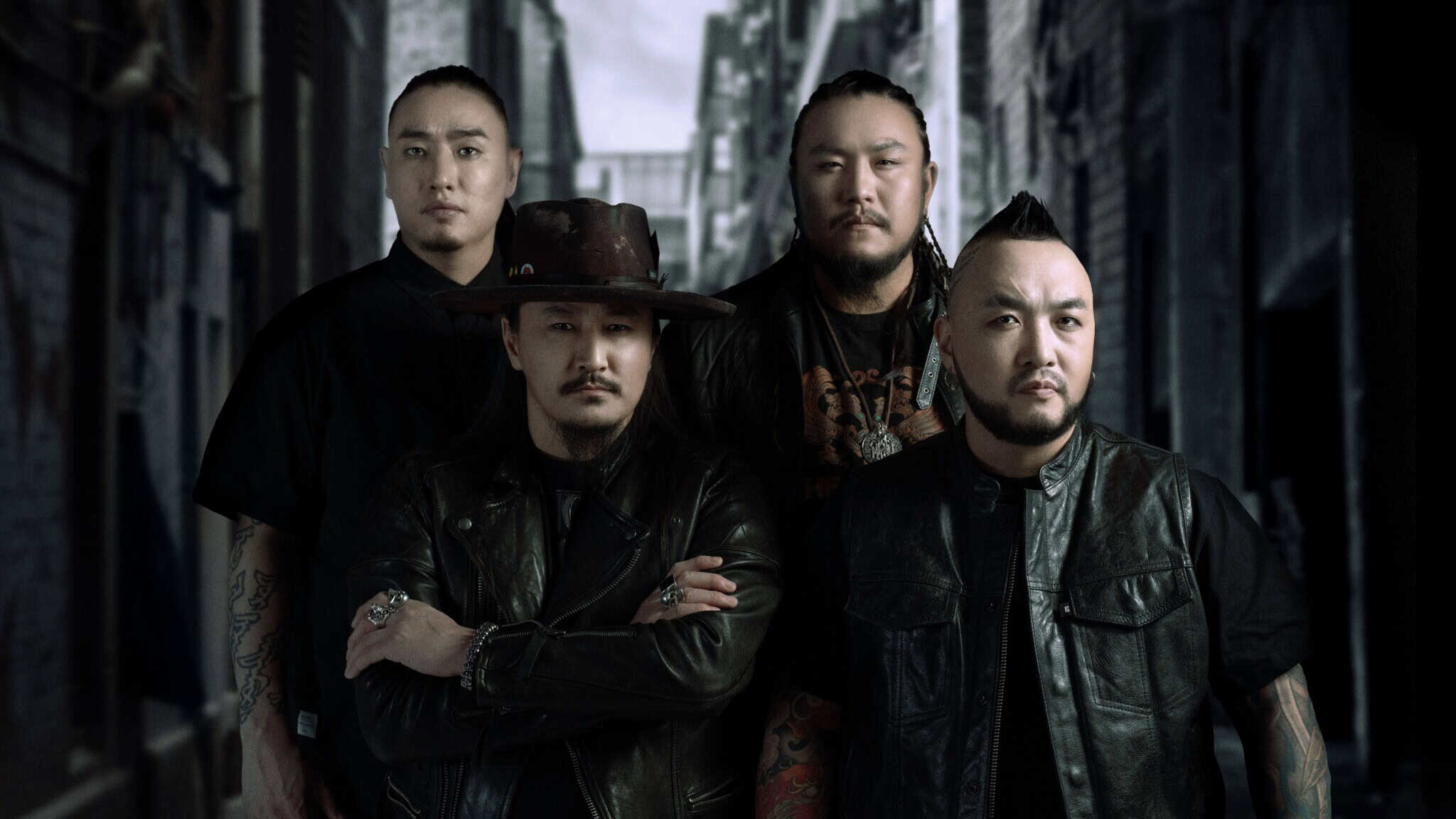 Four men stand together in a dark alleyway, all wearing black clothing and serious expressions. One man in front wears a wide-brimmed hat and crosses his arms, while the others stand closely behind him.