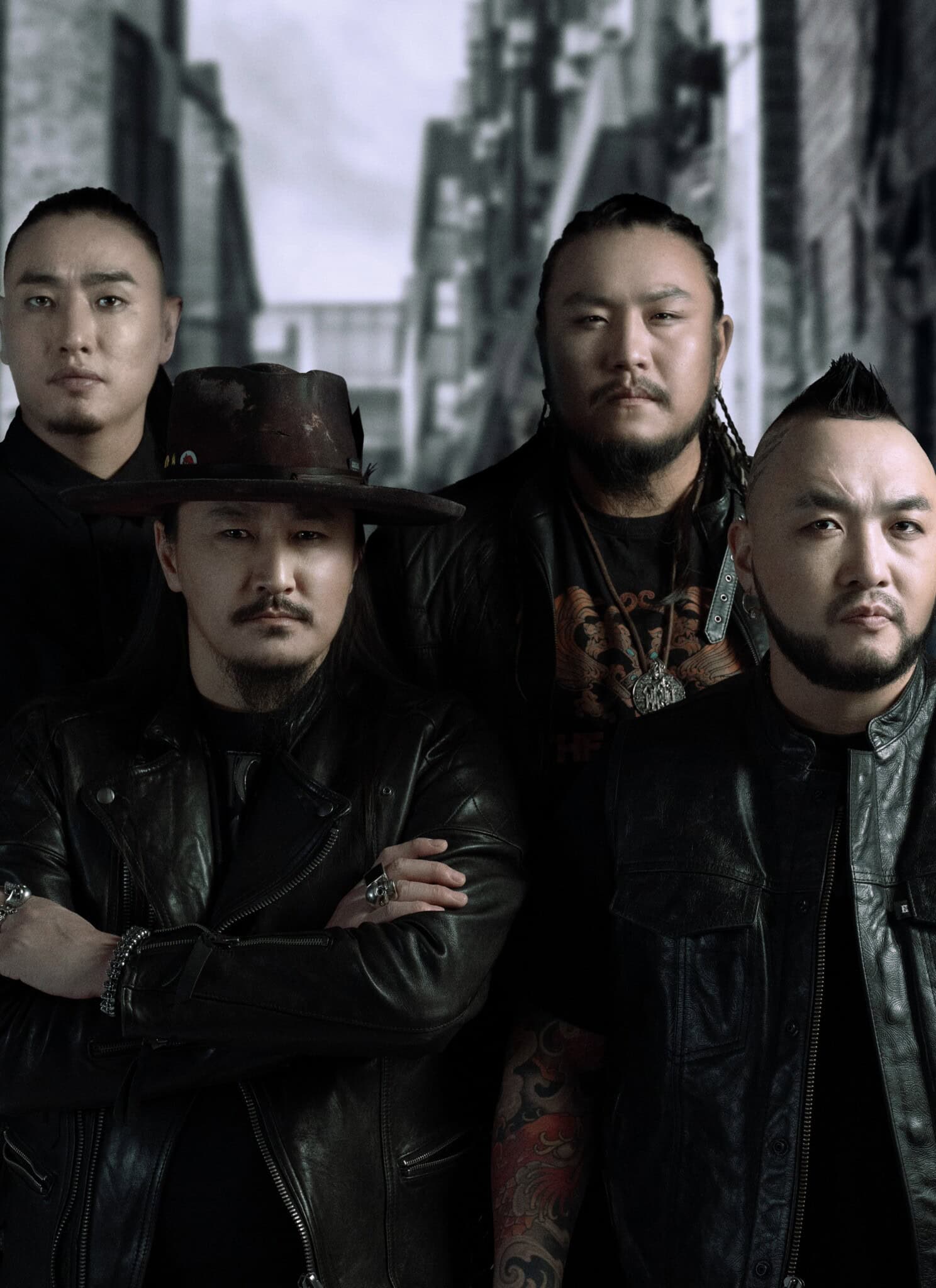 Four men stand together in a dark alleyway, all wearing black clothing and serious expressions. One man in front wears a wide-brimmed hat and crosses his arms, while the others stand closely behind him.