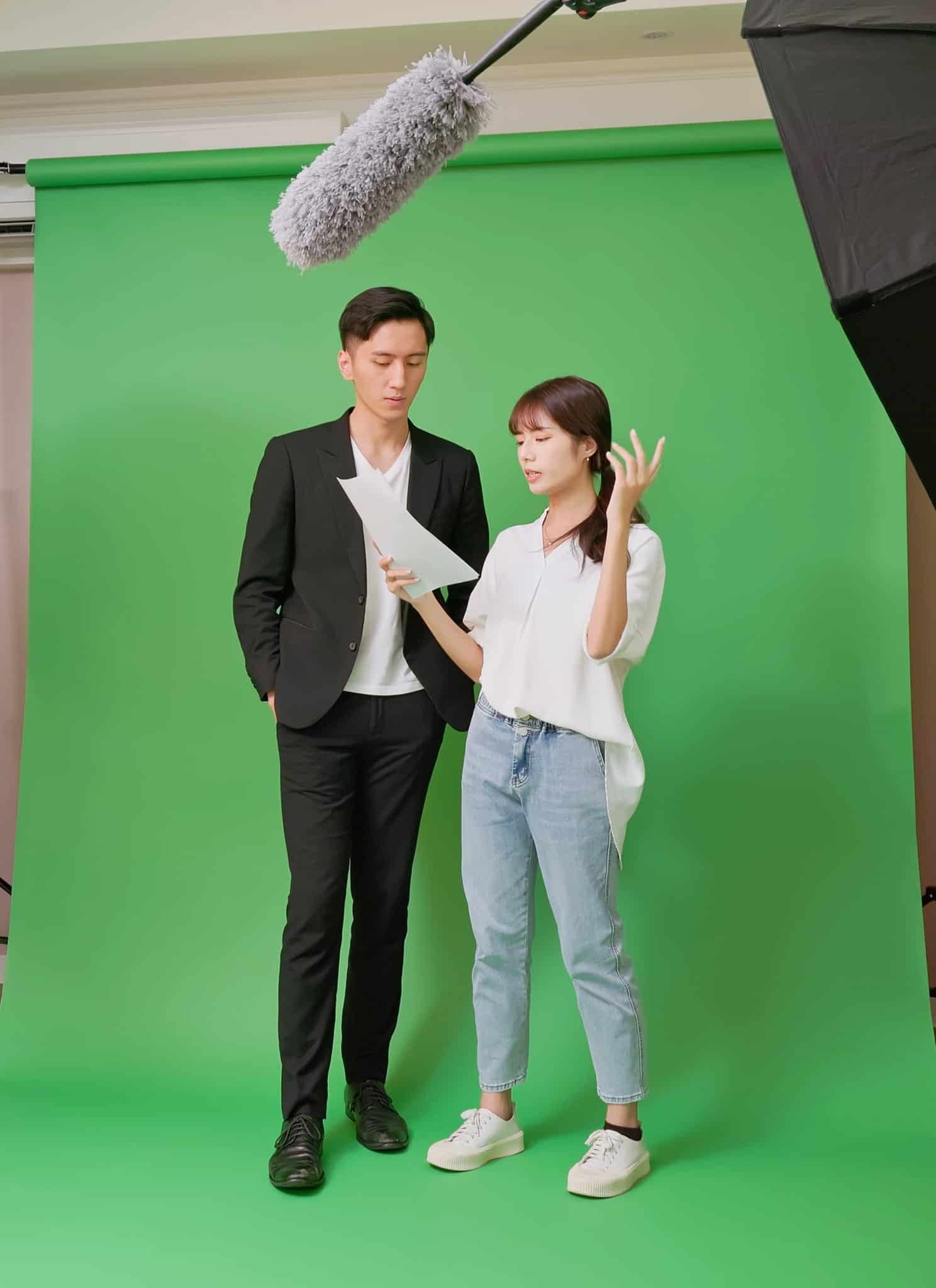 Two people stand in front of a green screen, discussing a script. Professional lighting, a camera, and a boom microphone are set up around them, with crew members adjusting equipment in a studio setting.