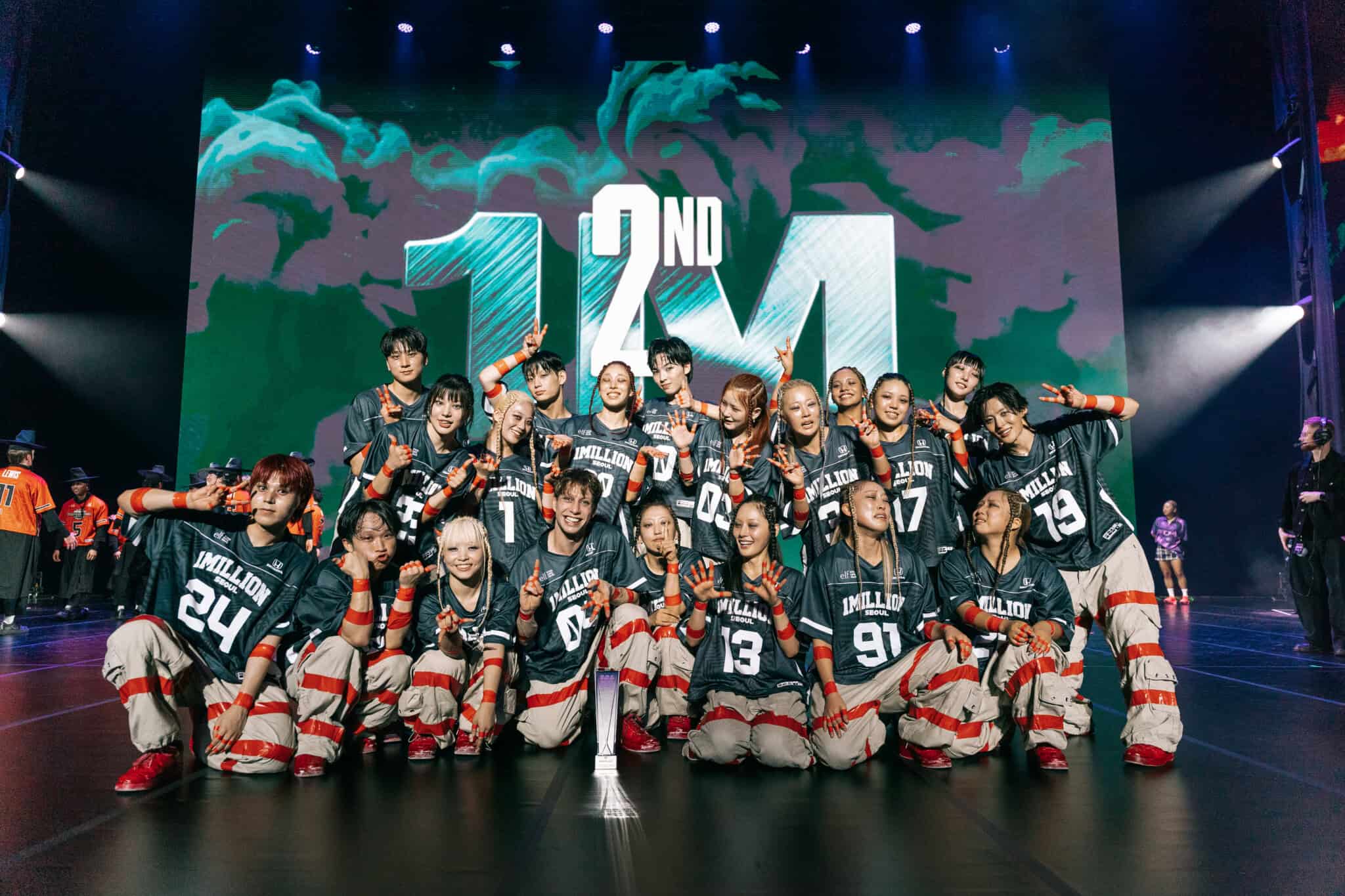 A large dance crew in matching uniforms poses on stage with a trophy, smiling and making peace signs. The background displays 2ND 1M in large text.