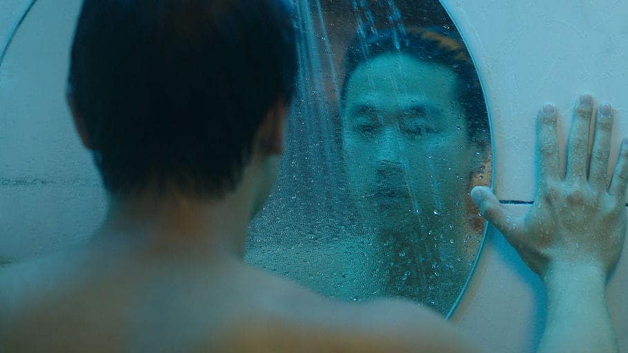 A person stands in front of a round mirror, touching it with one hand, while water droplets cover the glass. Their reflection appears blurred, and the scene has a cool blue tone.
