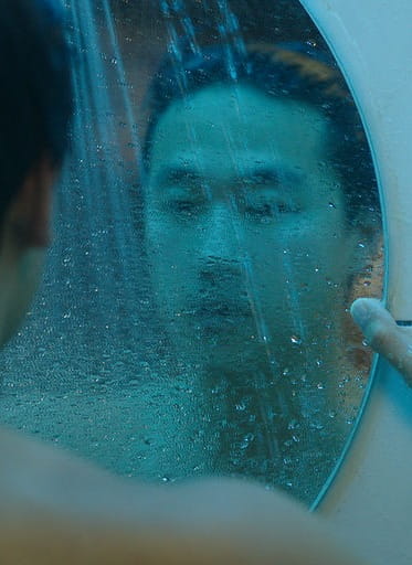 A person stands in front of a round mirror, touching it with one hand, while water droplets cover the glass. Their reflection appears blurred, and the scene has a cool blue tone.