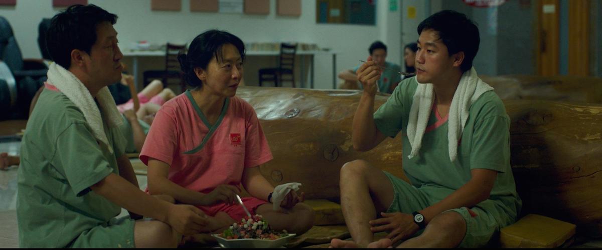 Three people in matching spa outfits sit on wooden benches, eating and talking in a relaxed indoor setting. Towels rest around their necks, and food is placed in front of them as they share a meal and conversation.