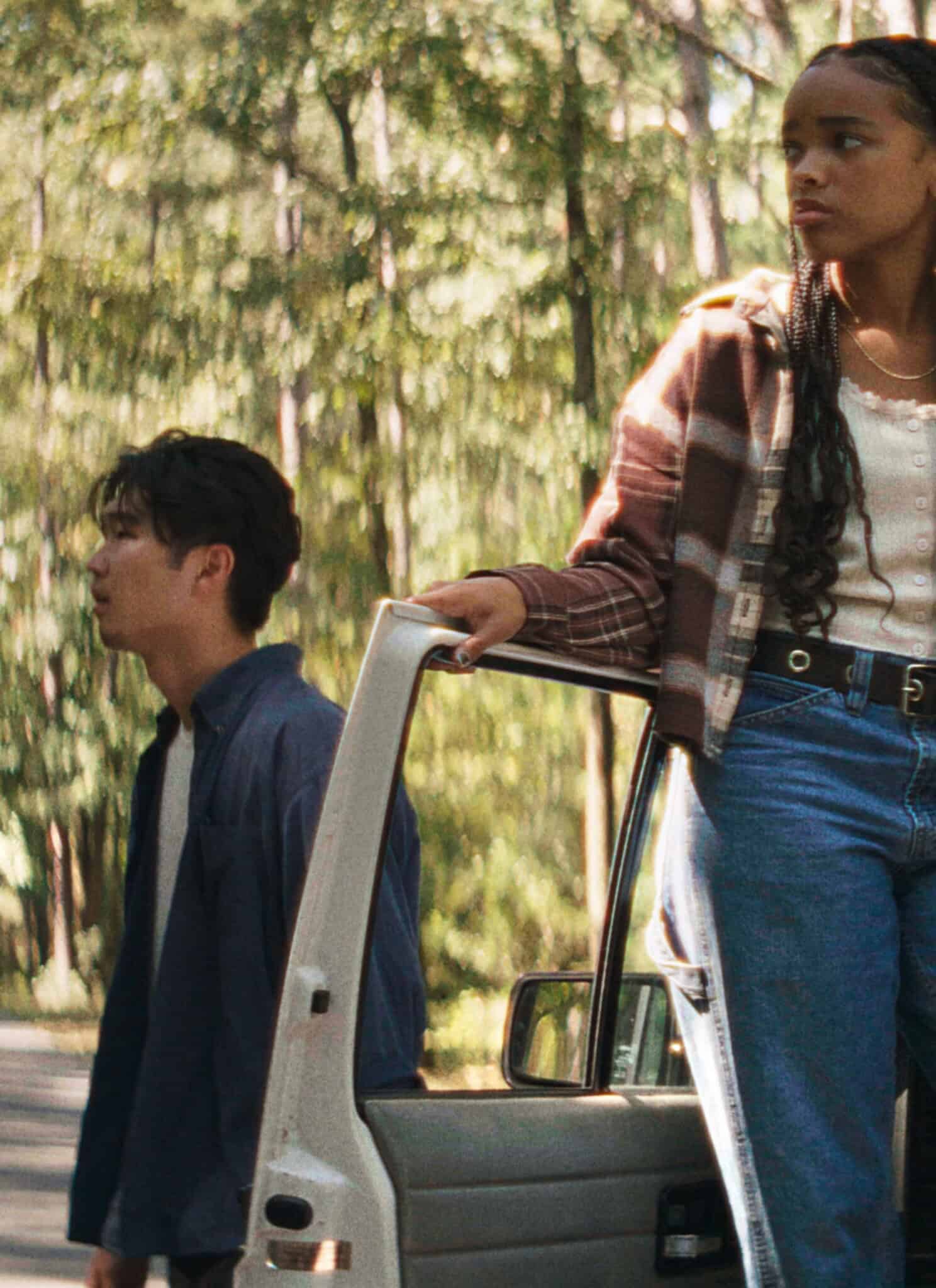 Three teenagers stand by a car stopped in a forested area. One girl leans out of the open car door, looking ahead, while two boys stand or sit nearby, all appearing concerned or thoughtful.