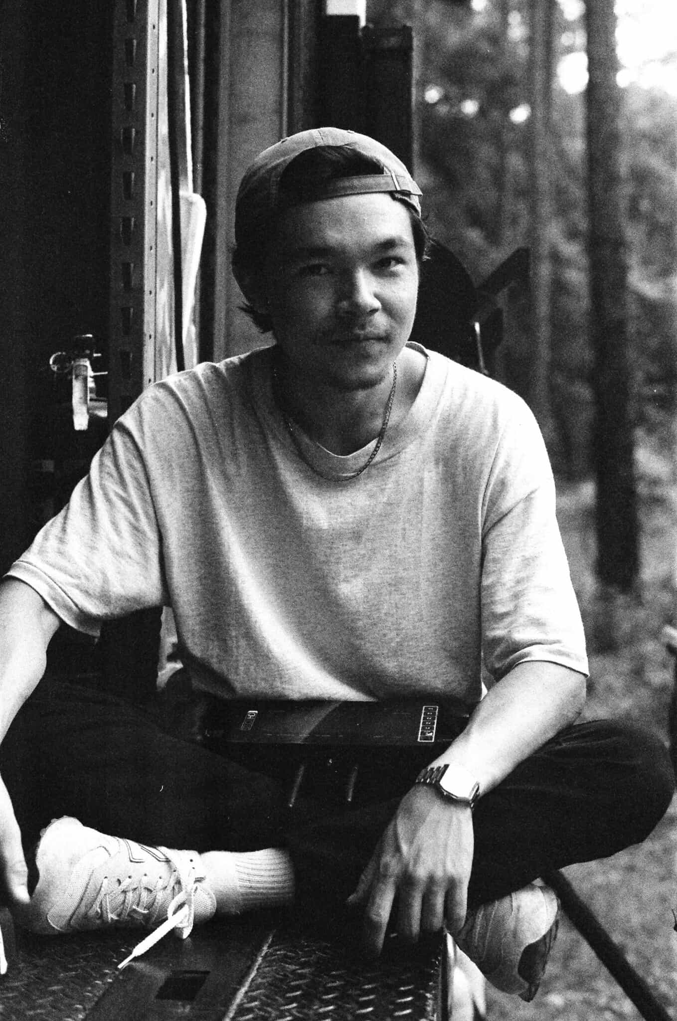 Black-and-white photo of a person sitting cross-legged outdoors, wearing a light shirt, dark pants, sneakers, and a backwards cap, looking at the camera and smiling softly.