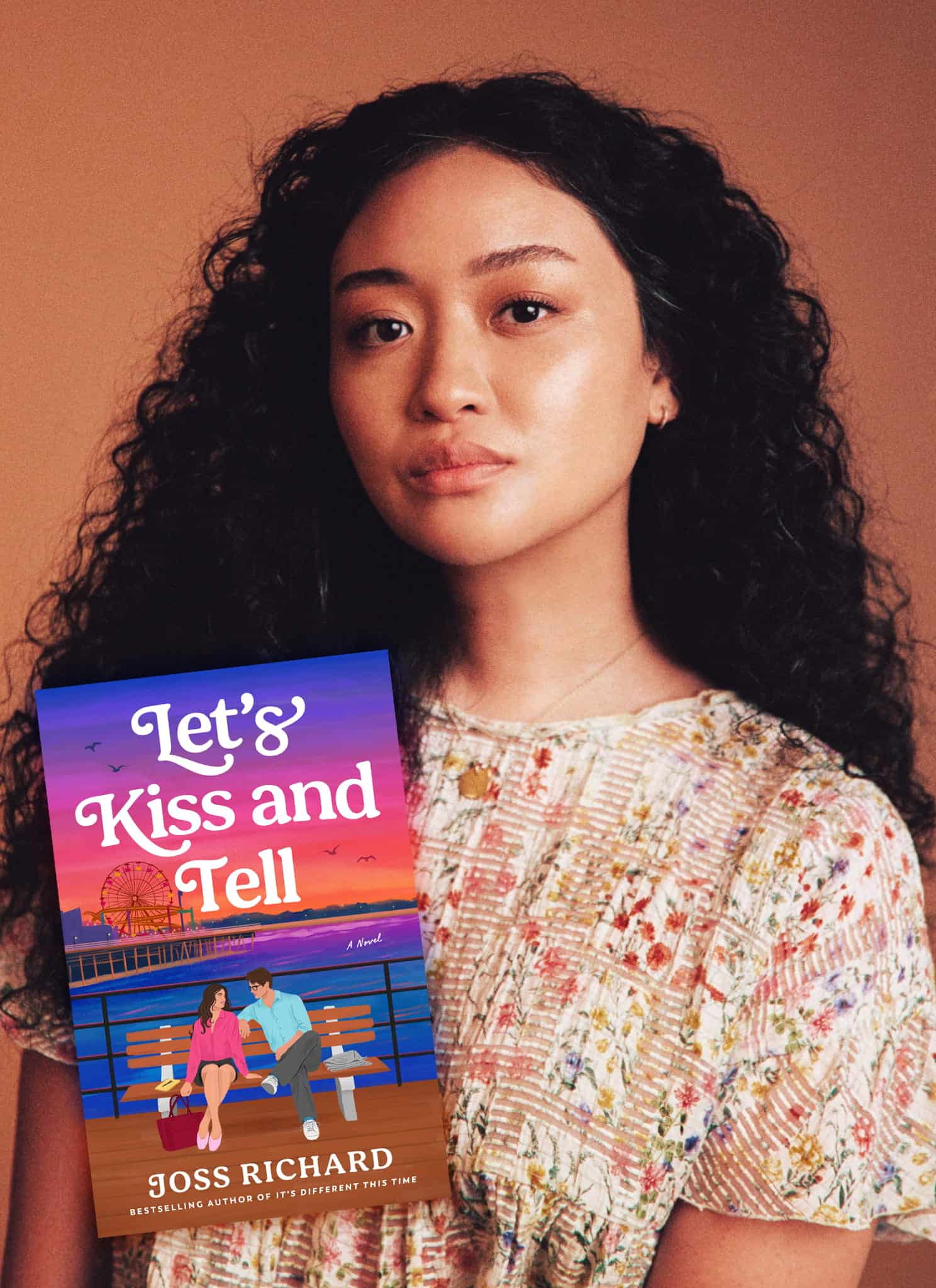 A woman with long curly hair stands next to a brightly colored book cover titled Lets Kiss and Tell by Joss Richard, showing a couple sitting on a bench by a pier at sunset.