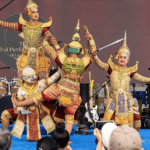 Four performers in ornate traditional Thai costumes enact a dramatic scene on stage, with two holding swords and others in theatrical poses. Musical instruments and audience members are visible in the background.