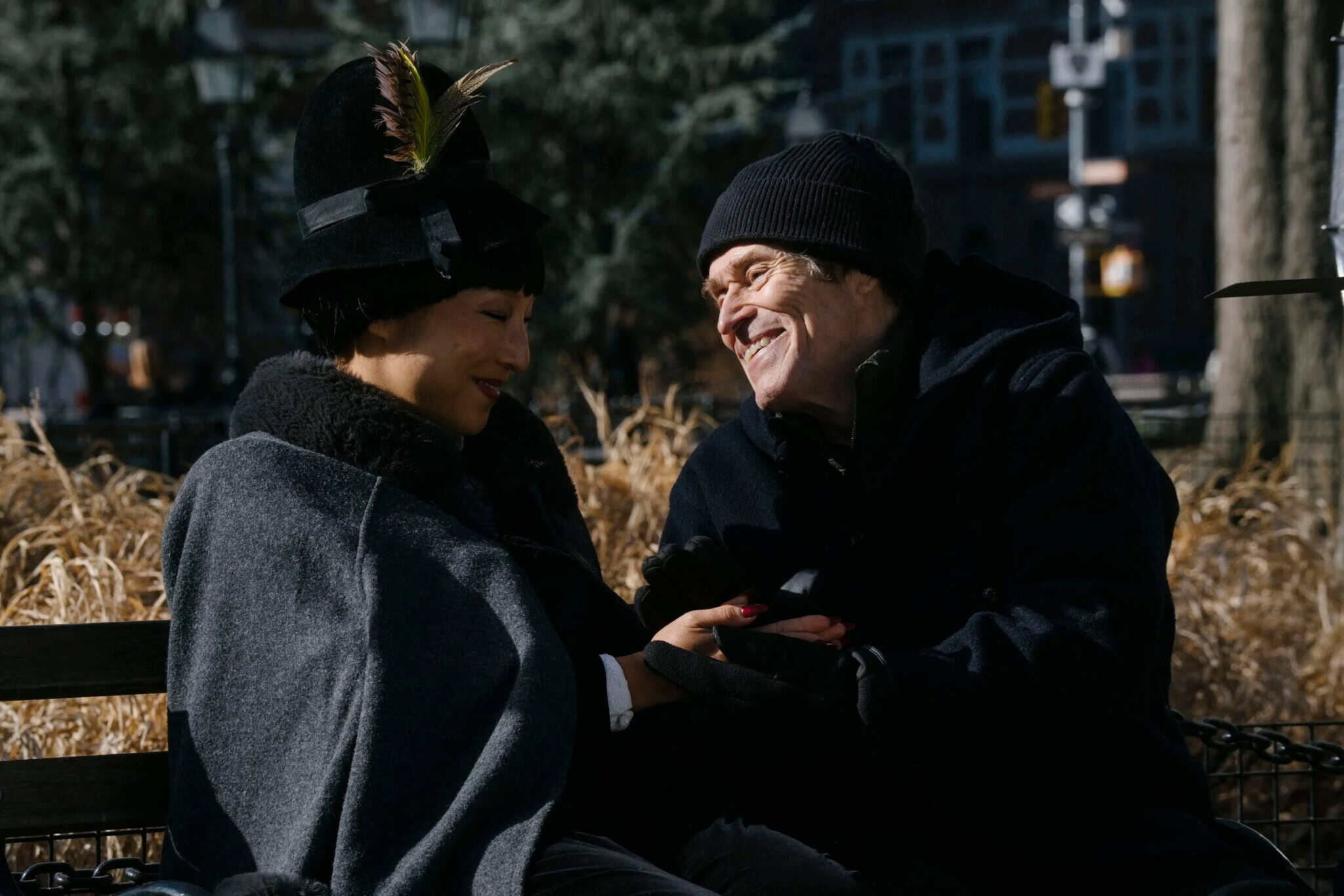 Two people sit close together on a park bench, smiling and holding hands. They are wearing warm coats and hats, with dry grass and trees in the background, suggesting a chilly day.