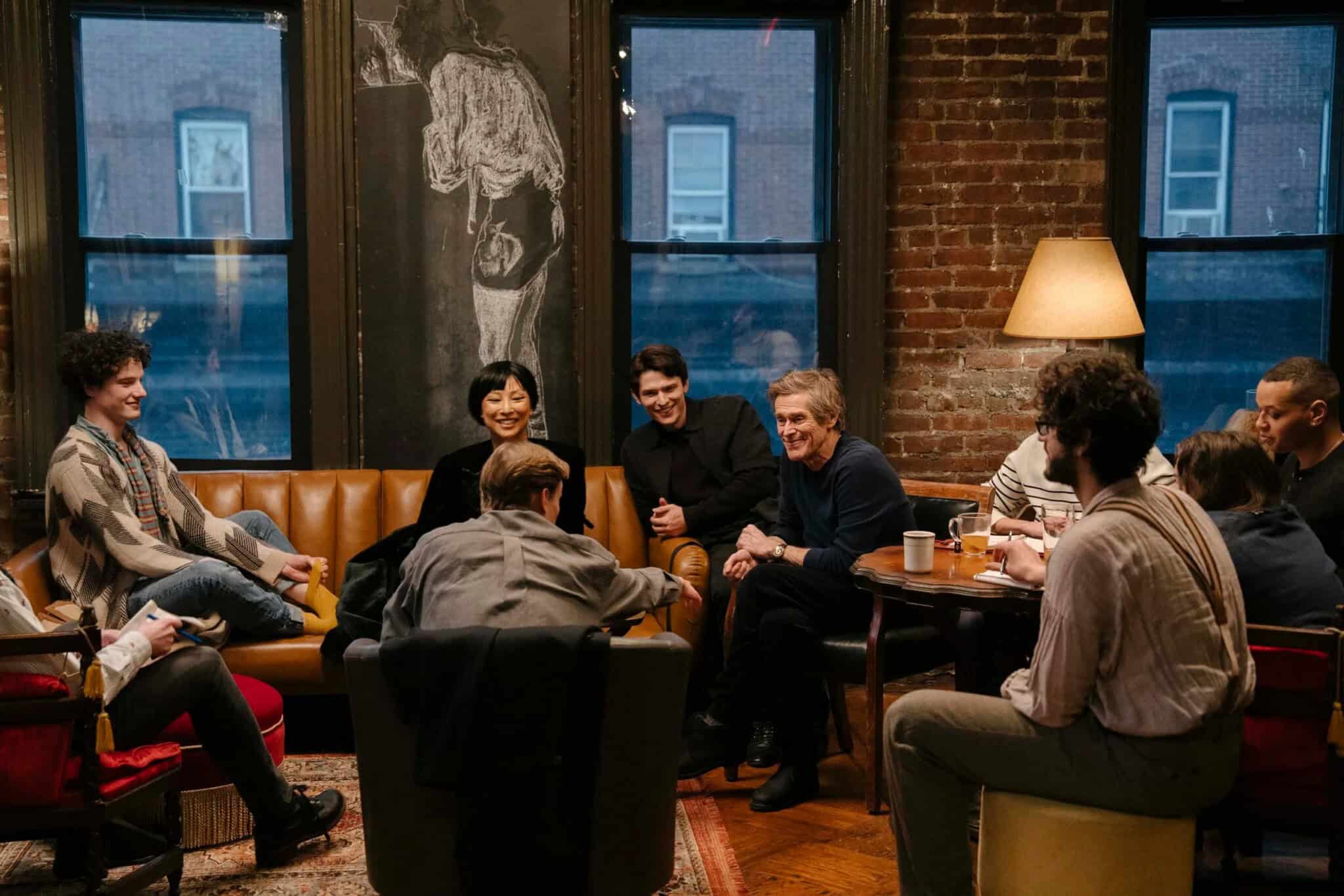 A group of people sit and talk around a coffee table in a cozy room with brick walls, large windows, and warm lighting.