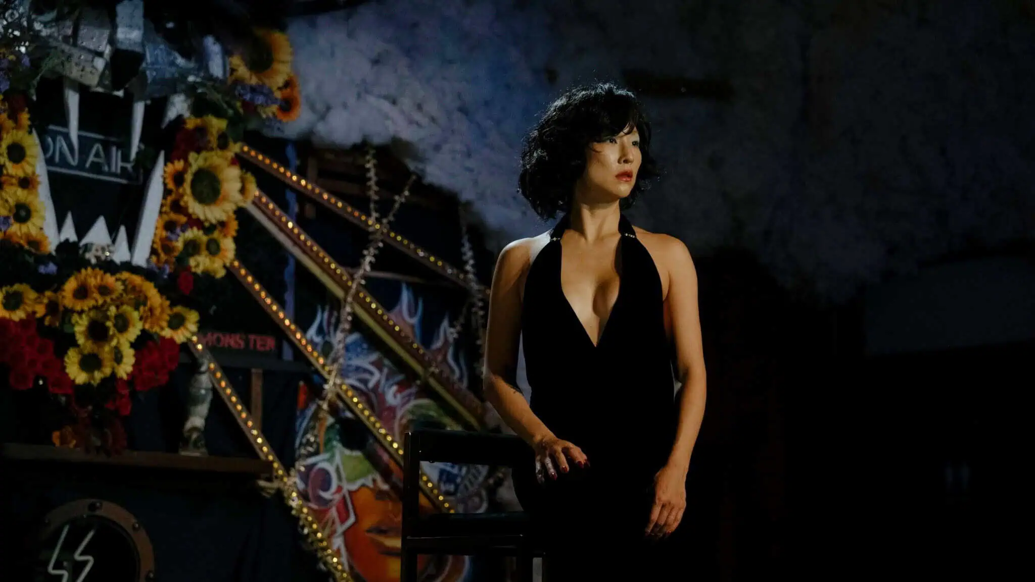 A woman in a black dress stands beside a chair on a dimly lit stage decorated with sunflowers, lights, and colorful graffiti-style art.