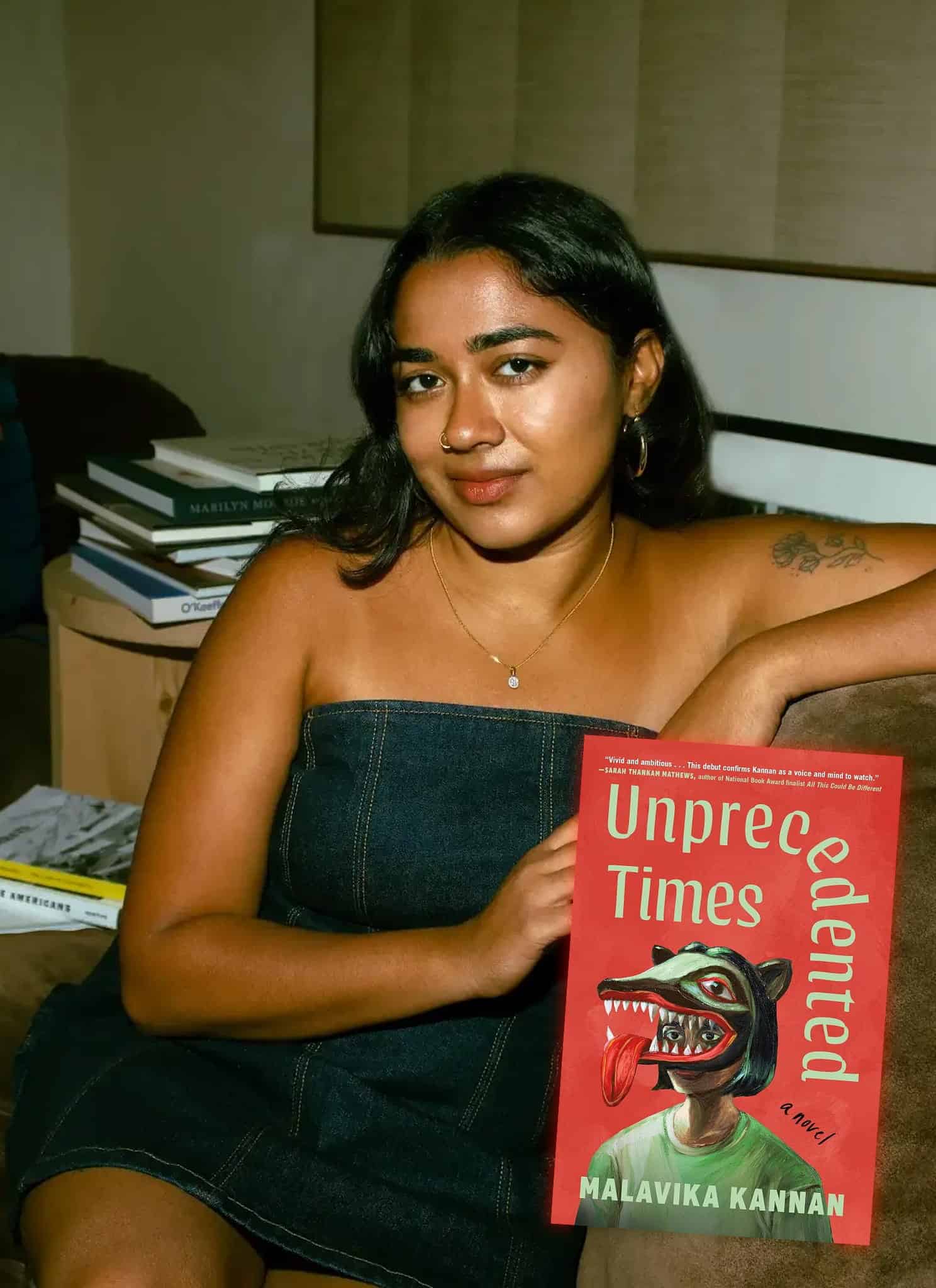 A woman sits beside a stack of books, next to the cover of the novel Unprecedented Times by Malavika Kannan, which features a stylized illustration of a person wearing an animal mask.
