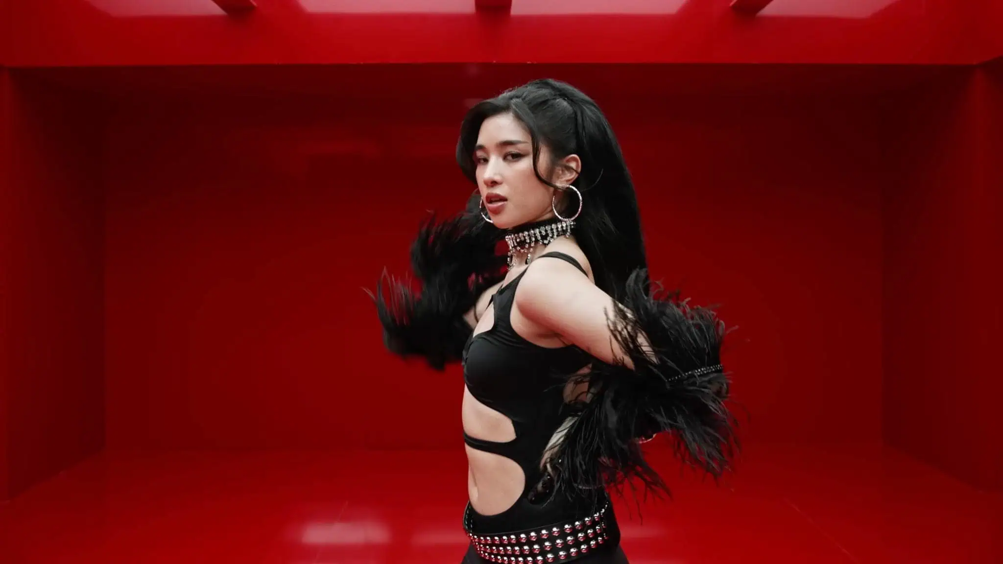 A woman with long dark hair poses confidently in a red room, wearing a black outfit with feathered sleeves, large hoop earrings, and a studded belt.