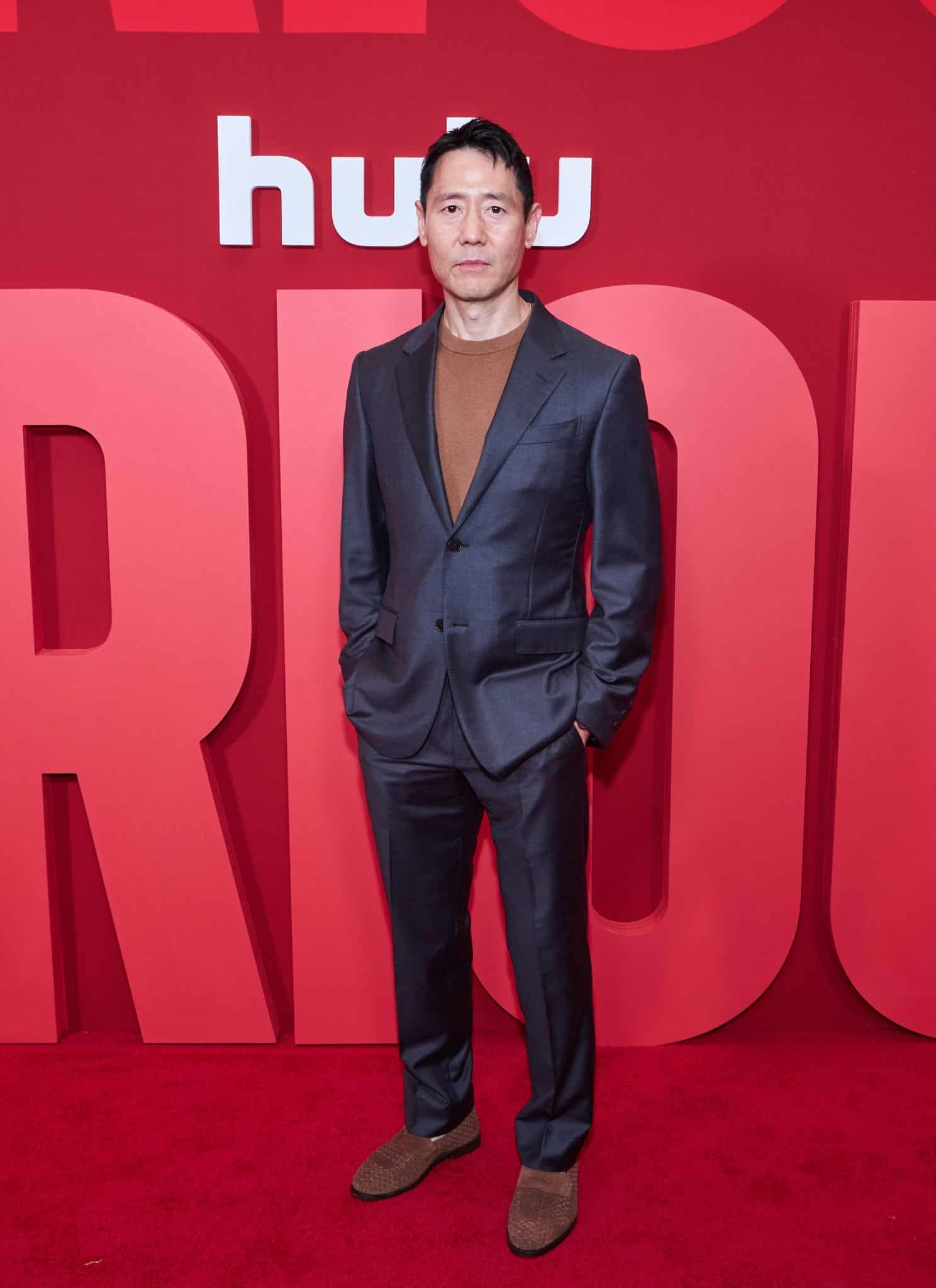 Rob Yang, dressed in a gray suit and brown shirt, stands in front of a red backdrop adorned with the white Hulu logo.